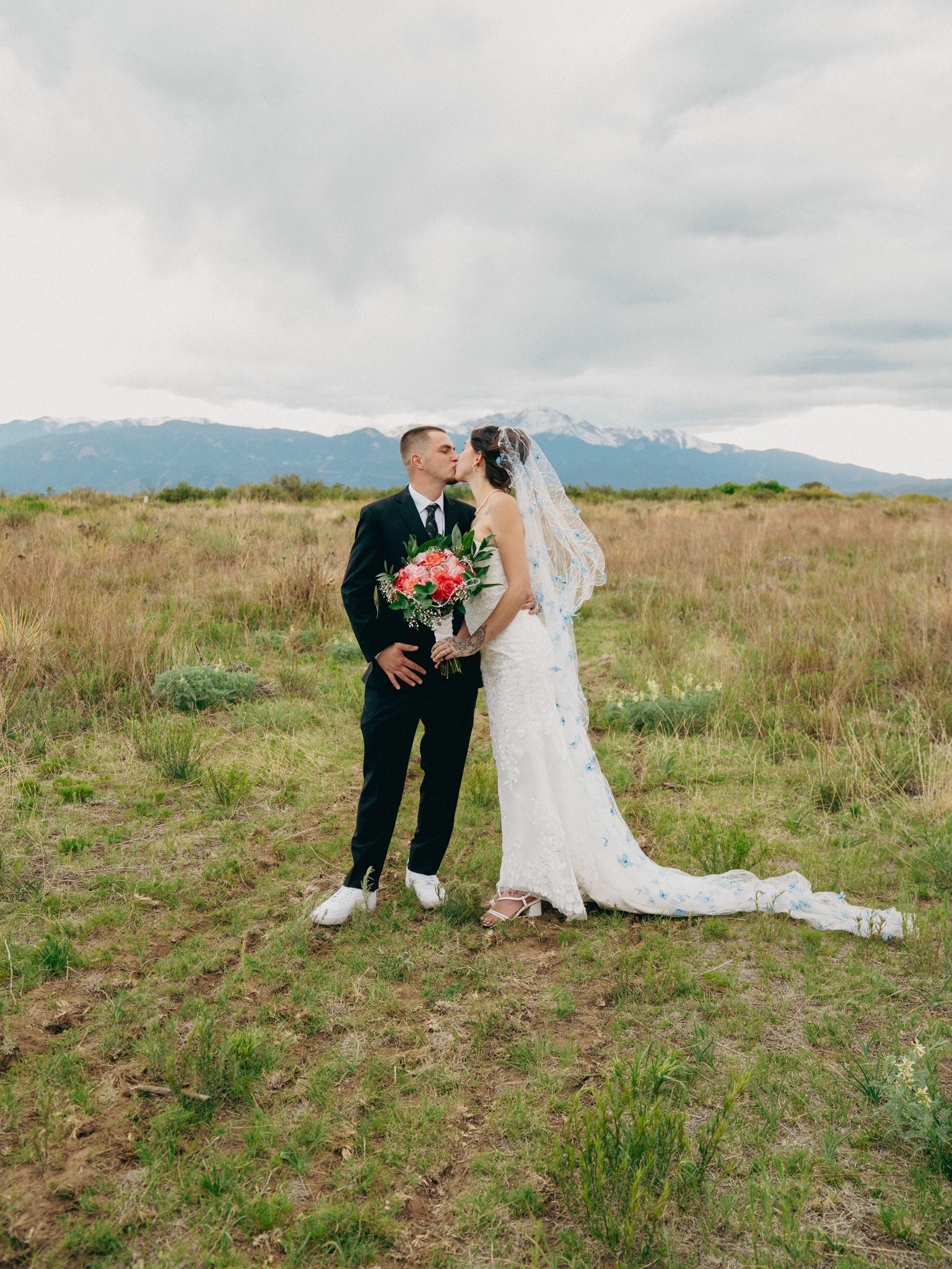 A couple in wedding attire sharing a kiss in an open grassy field with mountains in the background.