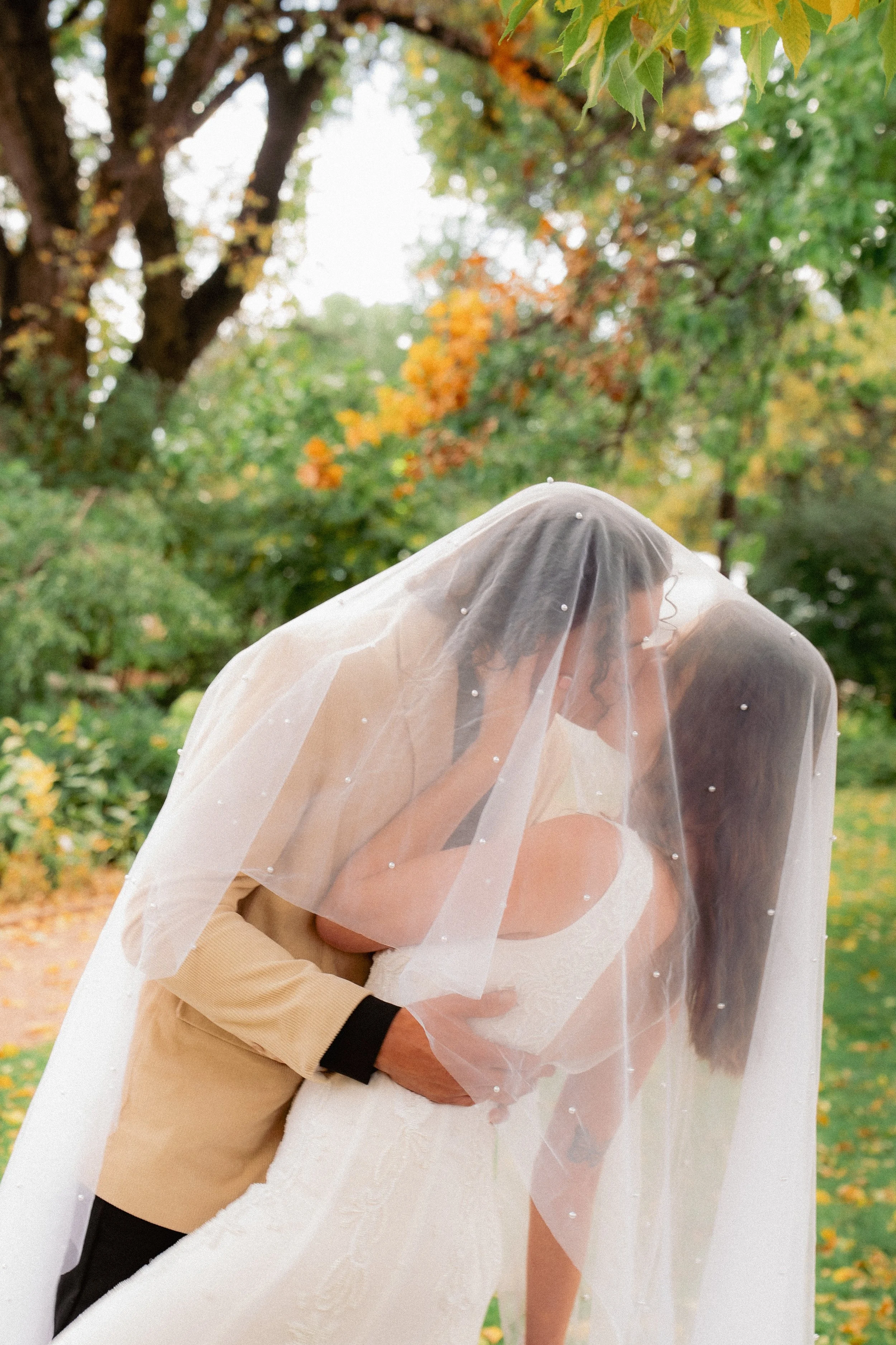 A bride and groom sharing a kiss under a veil on a sunny outdoor day, surrounded by green and autumn-colored foliage.