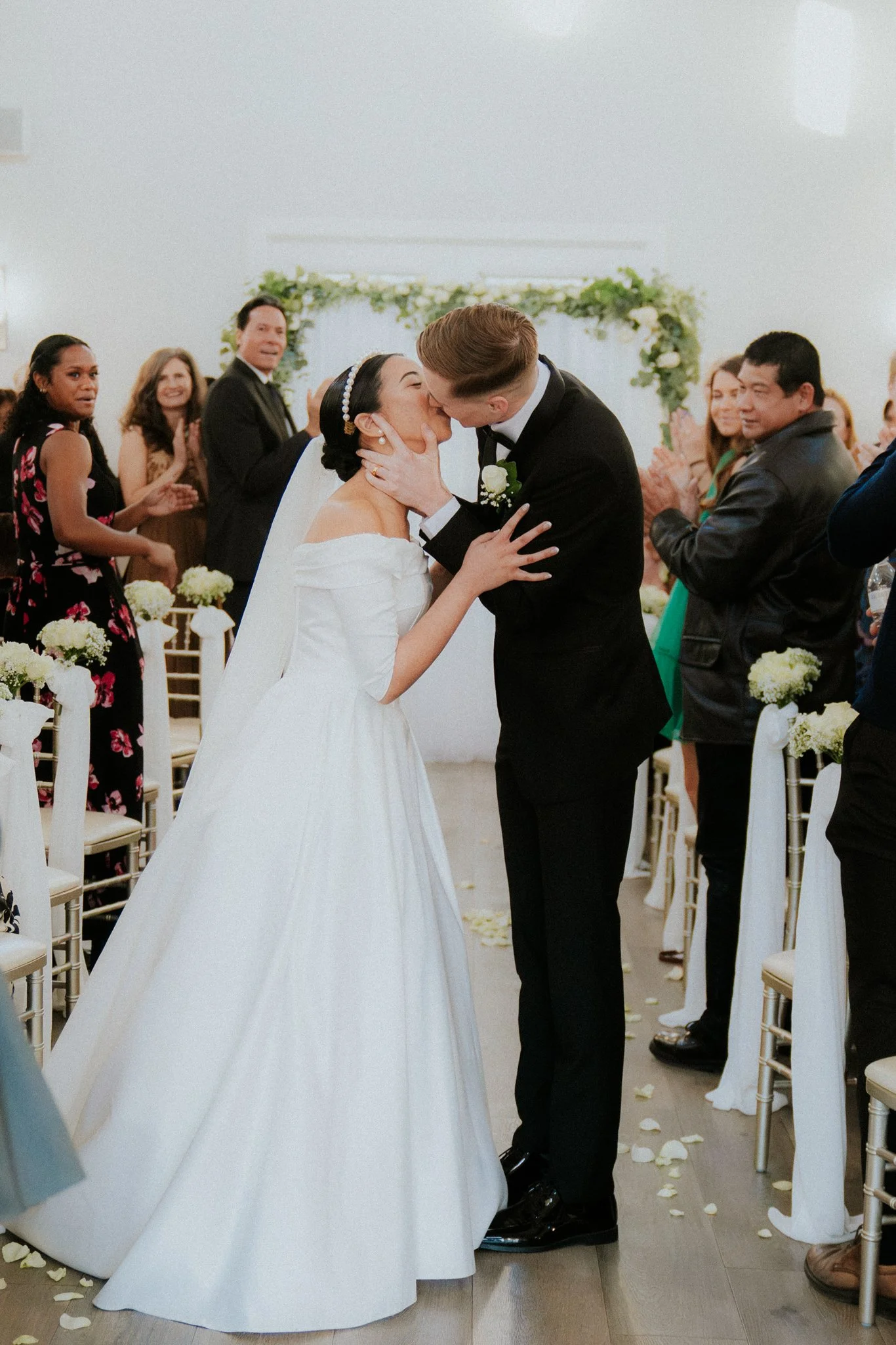A newlywed couple kissing during their wedding ceremony, surrounded by friends and family in a decorated venue.