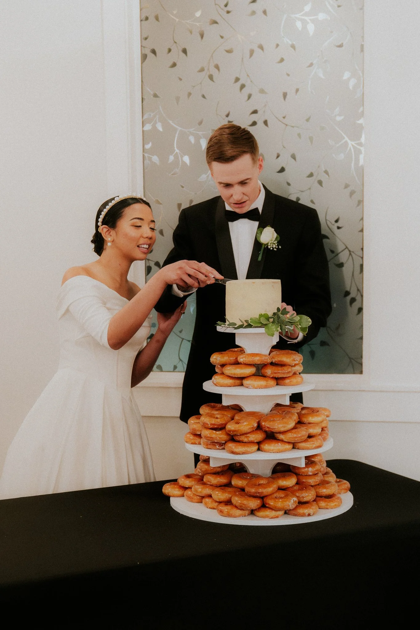 A bride and groom cutting a wedding cake together. The cake is small, square, with greenery and is on a stand surrounded by donuts.