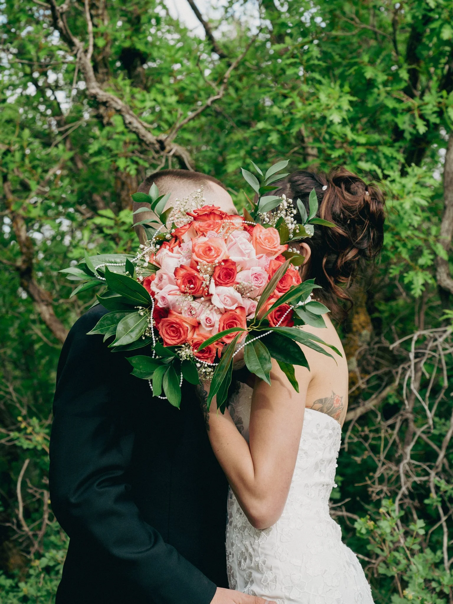 A newlywed couple is kissing outdoors, with the bride holding a bouquet of pink, peach, and red roses covering their faces. The groom is wearing a black suit, and the bride is in a white lace wedding dress with her hair styled in loose curls and a ta