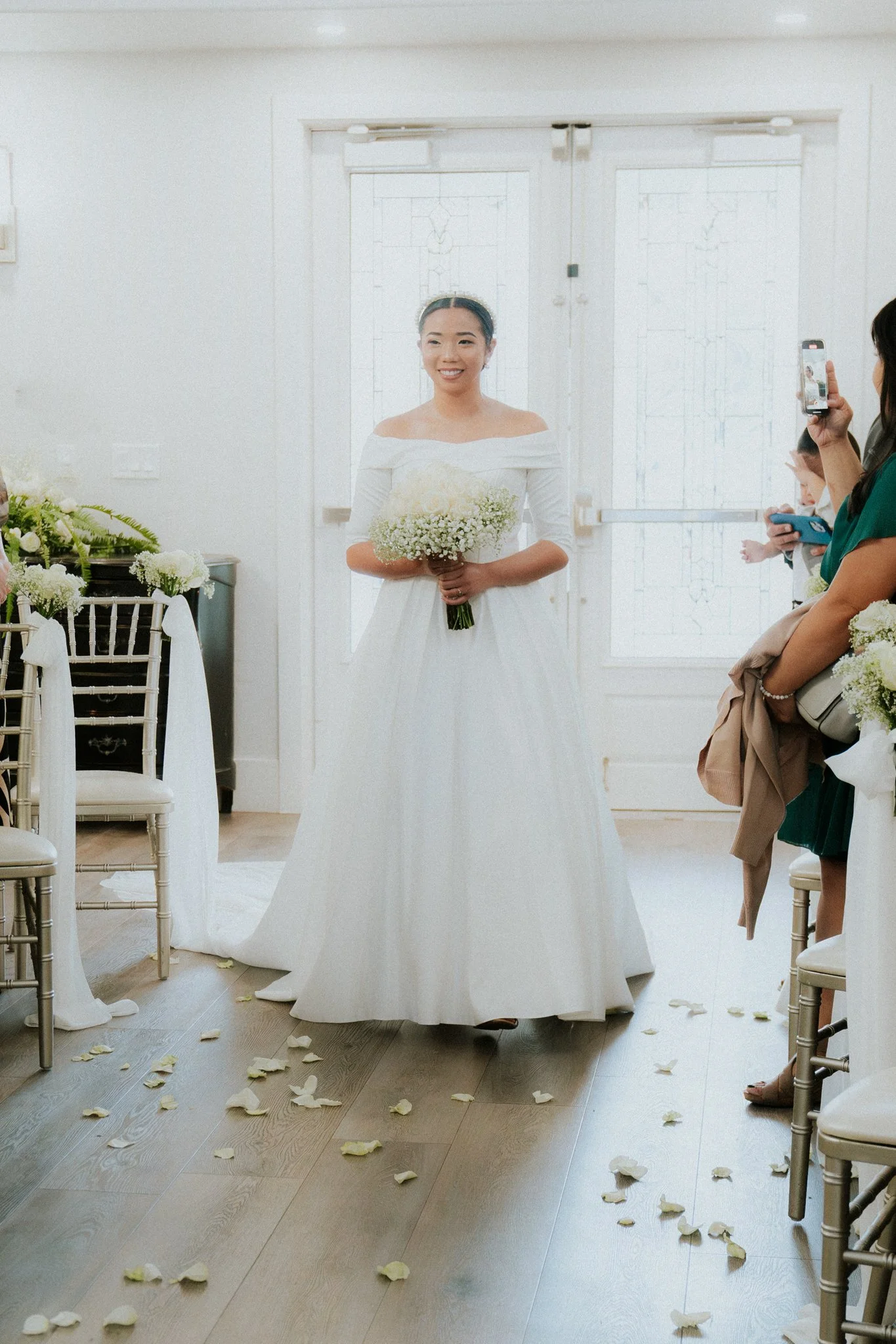 A bride in a white wedding dress holding a bouquet of flowers, standing in a decorated venue with guests taking pictures.