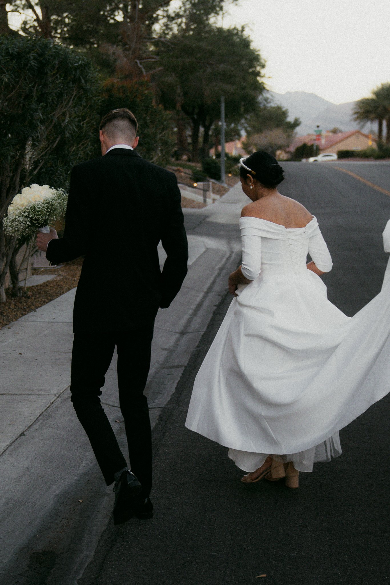 A bride and groom walking on a sidewalk in a suburban neighborhood during dusk, with the bride wearing a white off-the-shoulder wedding dress and the groom in a black suit holding a bouquet of flowers.