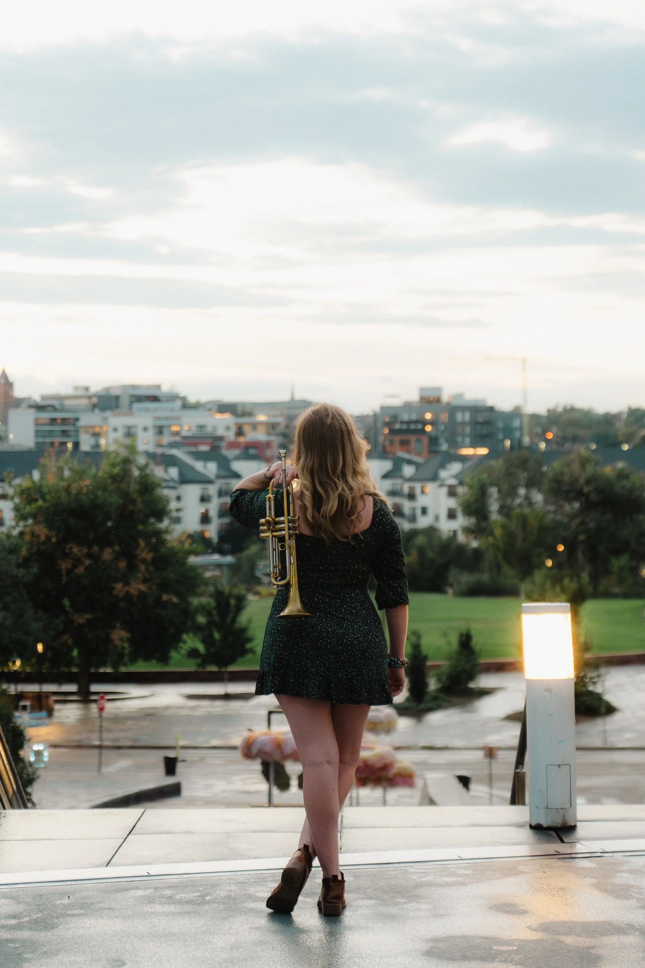A woman with wavy blonde hair, wearing a black dress, standing outdoors at dusk with a trumpet on her shoulder, looking at a cityscape with modern buildings and trees.