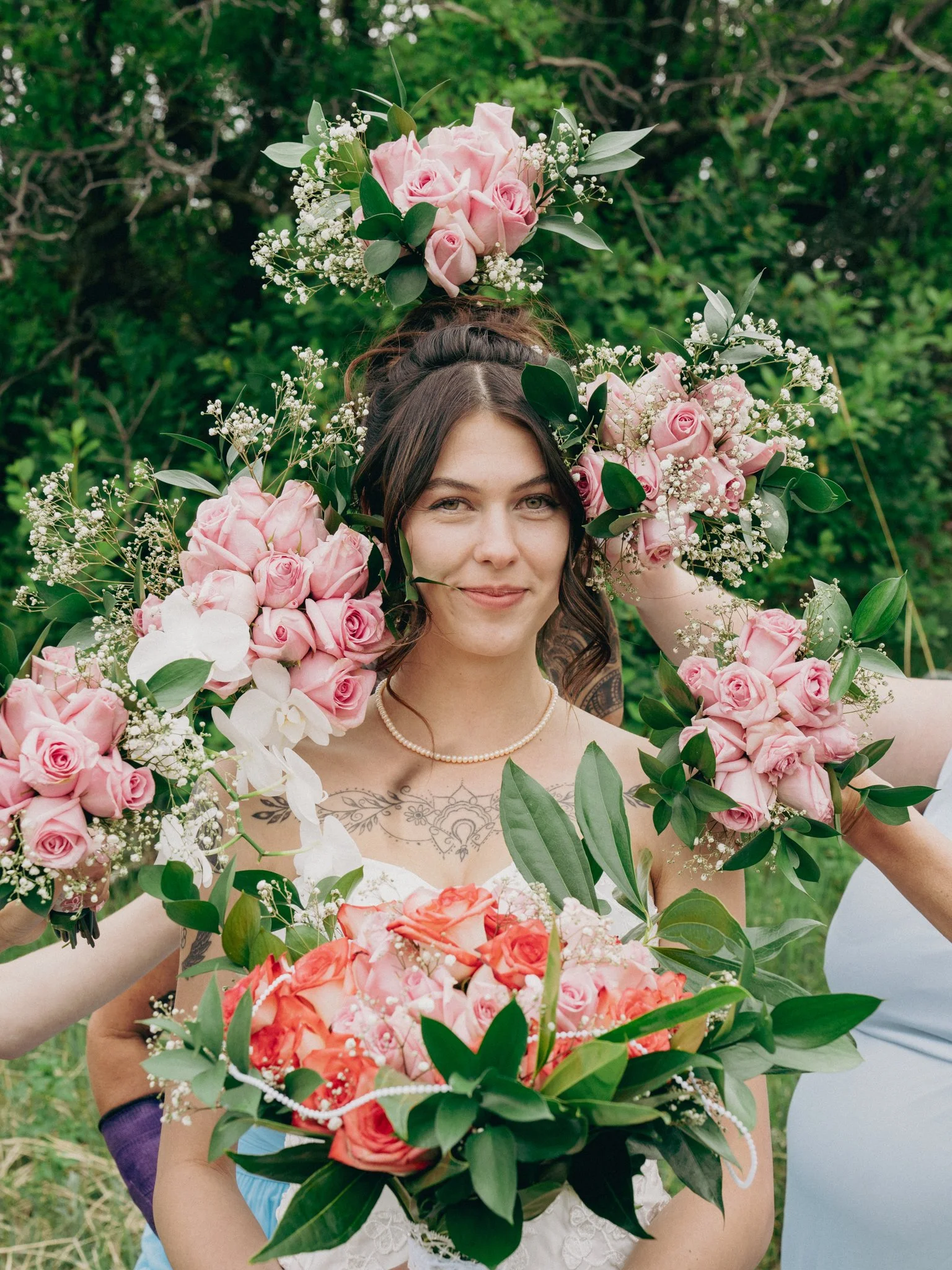 A woman with dark hair holding a large bouquet of pink and white roses, surrounded by greenery, during a wedding or celebration.