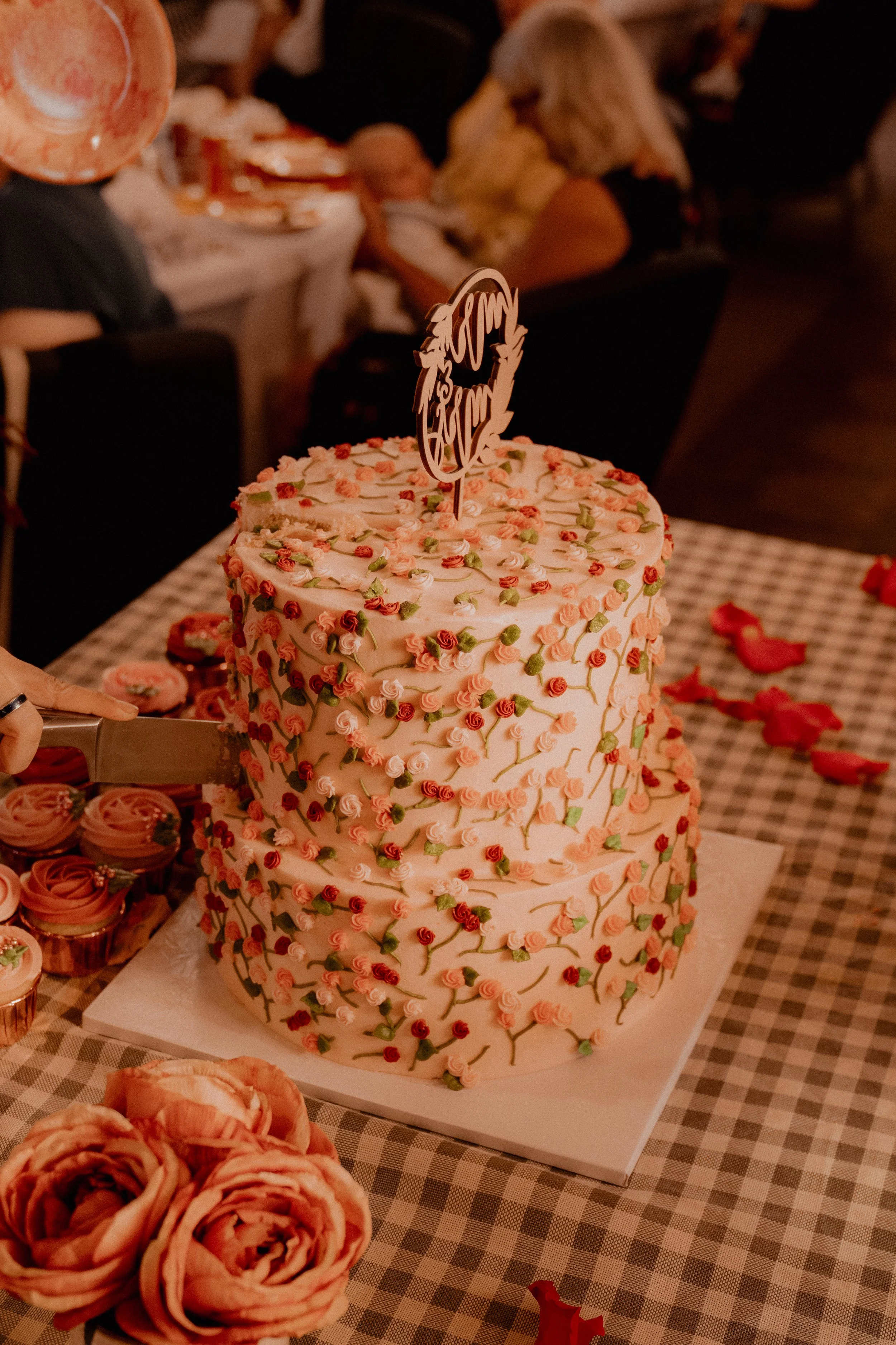 A three-tiered wedding cake decorated with pink, red, and green roses and leaves, with a topper that says 'Mrs. & Mrs.' on a chocolate stick, placed on a checkered tablecloth at a celebration event.