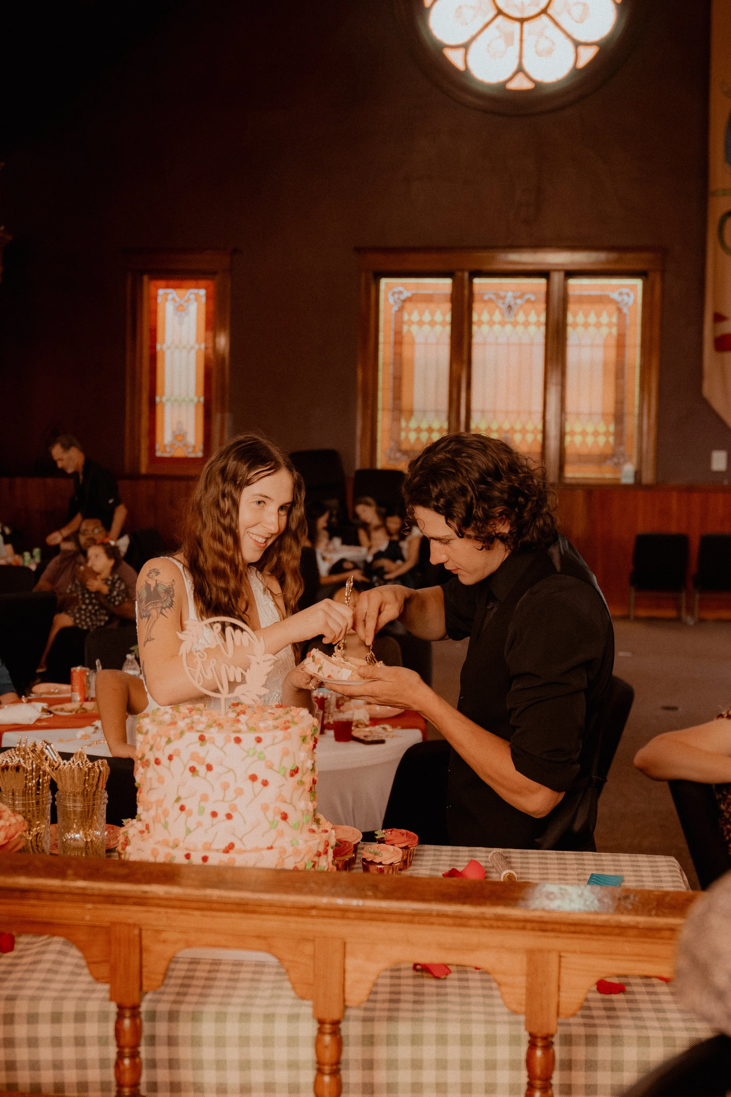 A birthday celebration with a girl smiling as she receives a gift, a large decorated cake on the table, and in the background, children and adults in a warmly lit room.