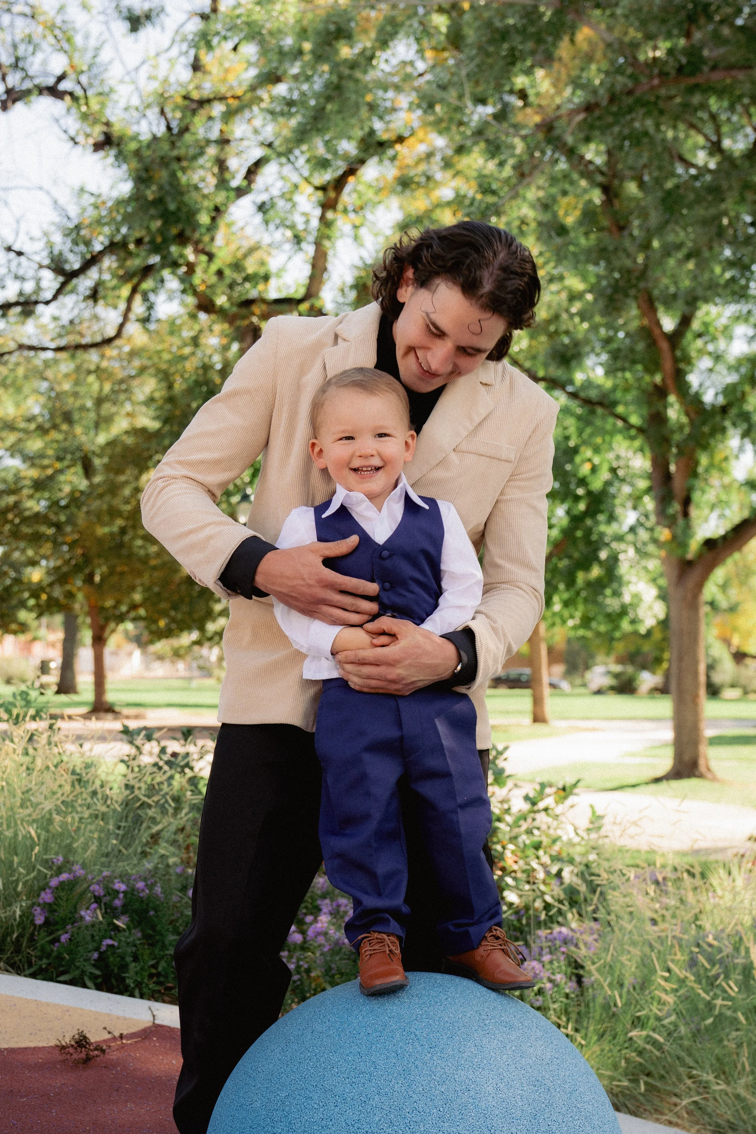 A man with curly dark hair and a beige jacket is holding a young smiling boy in a blue vest and pants, standing on a blue exercise ball at a park with green trees and purple flowers in the background.