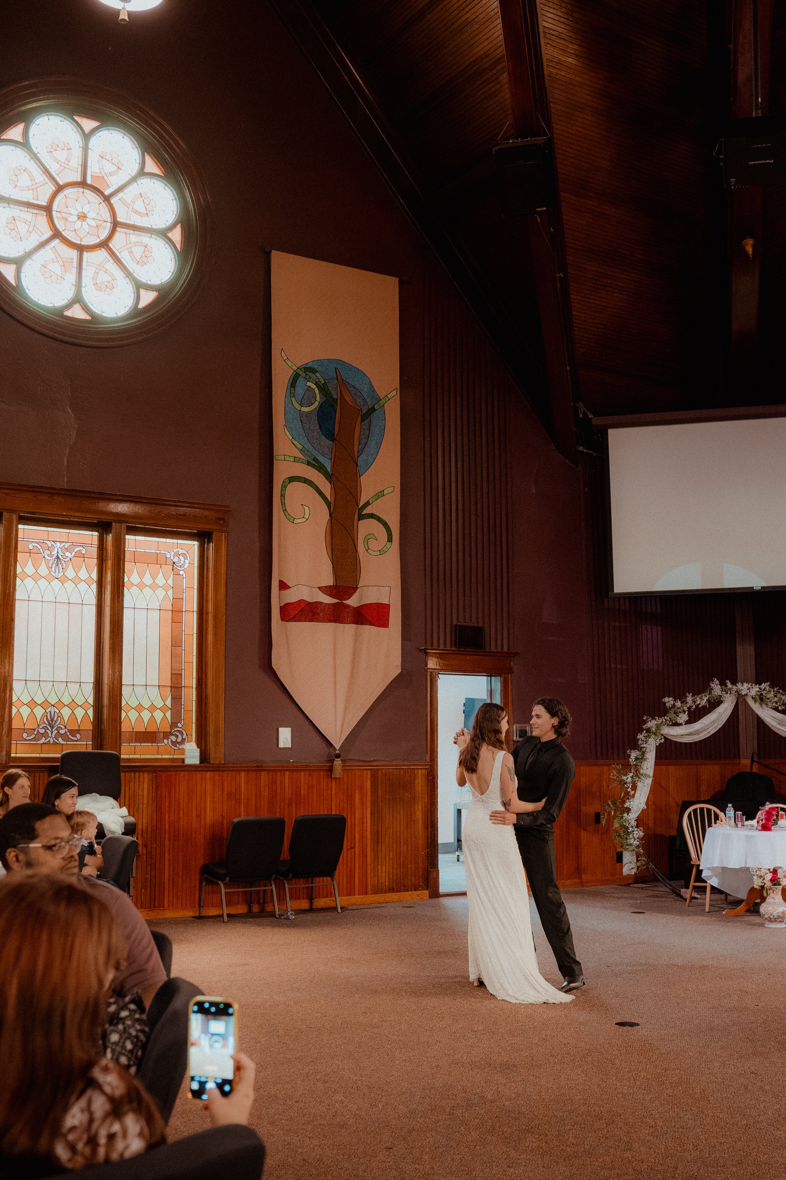 A bride and groom dancing during their wedding reception in a church hall, with guests seated around watching.