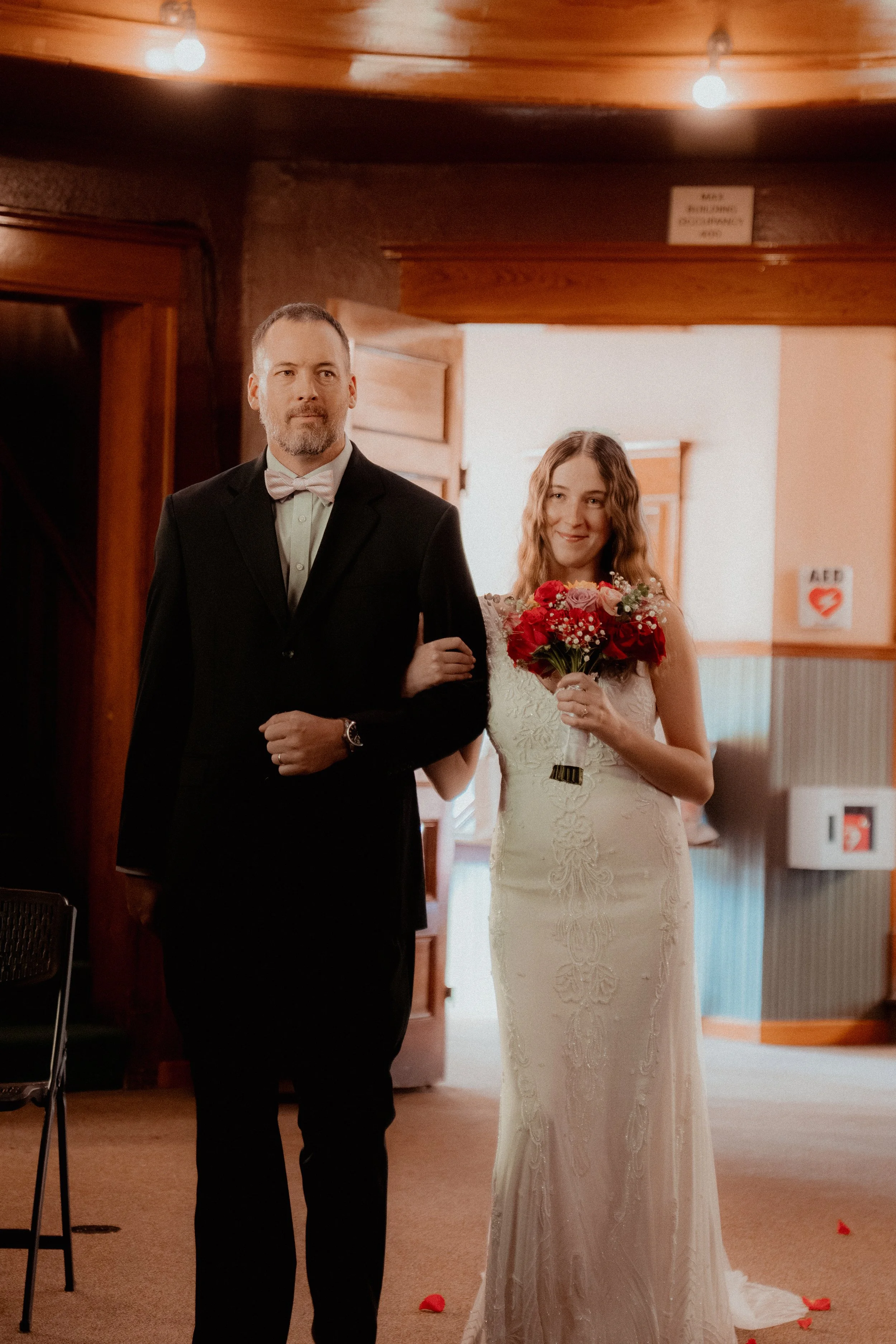 A groom in a black suit and white bow tie walking a bride with wavy hair holding a bouquet of red and pink flowers inside a wooden-paneled building.