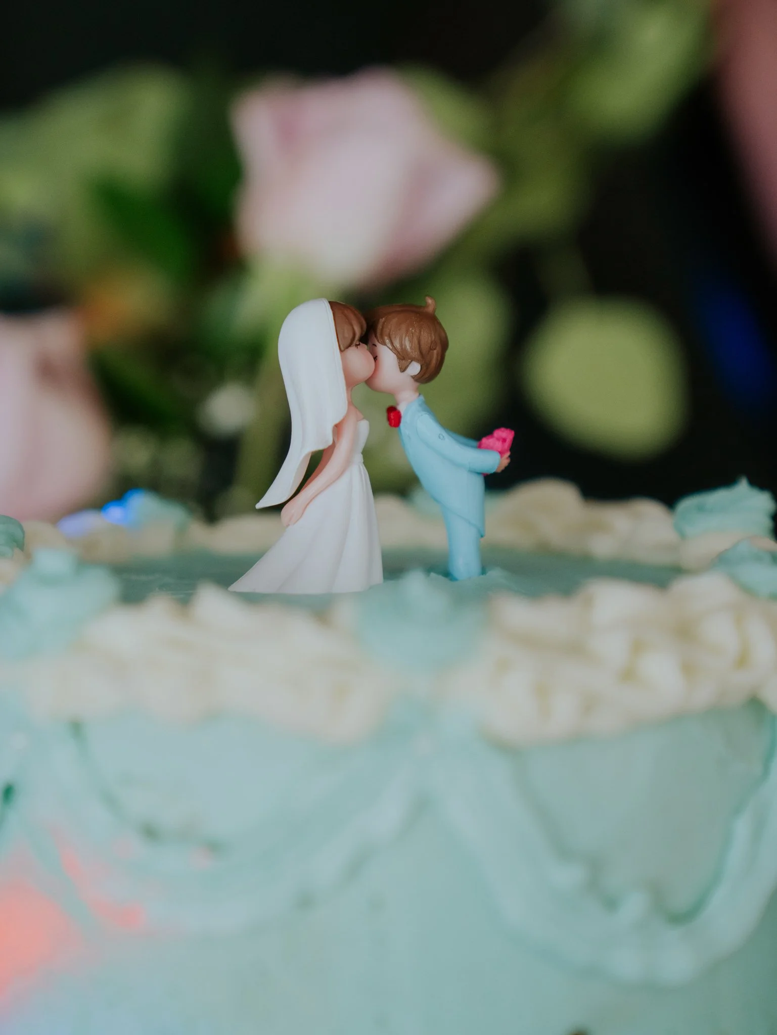 Cake topper featuring a bride and groom figurines, with their faces close together on top of a wedding cake decorated with icing.