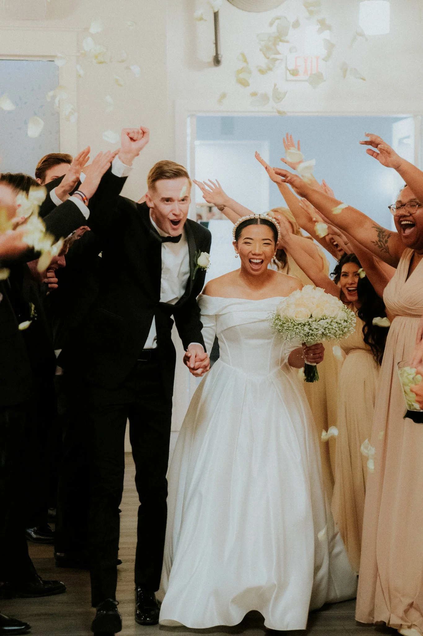 Bride and groom walking hand in hand through a celebration of friends and family, with guests throwing flower petals in the air.