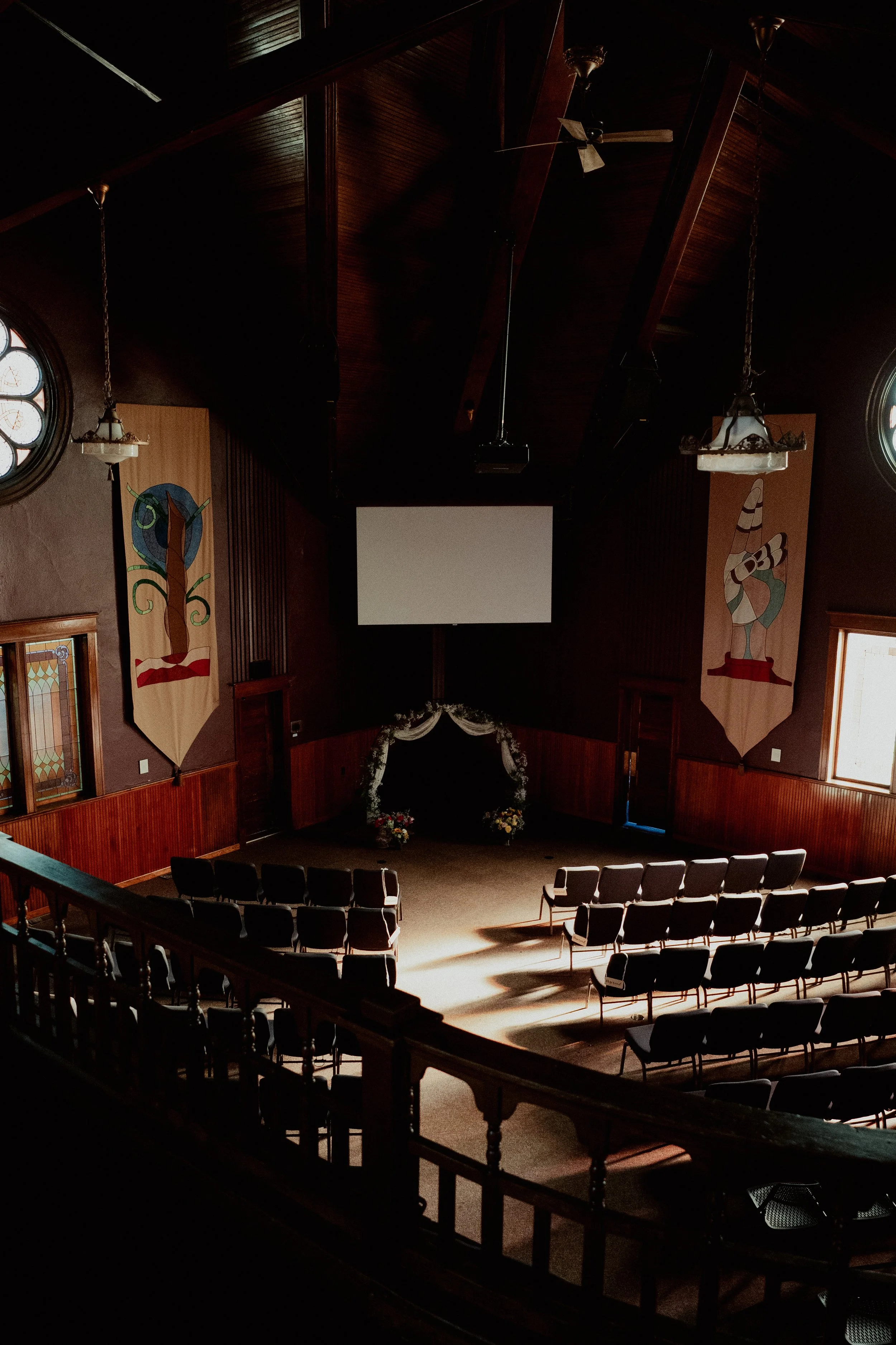 Empty chapel with rows of chairs facing a screen and a floral arch at the front, stained glass windows, wooden walls, and banners with artistic designs.