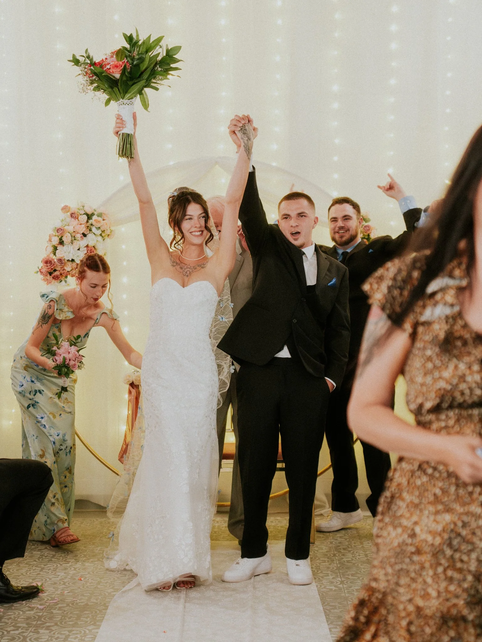 Bride and groom celebrating at their wedding reception, bride holding a bouquet with both hands raised, surrounded by guests.