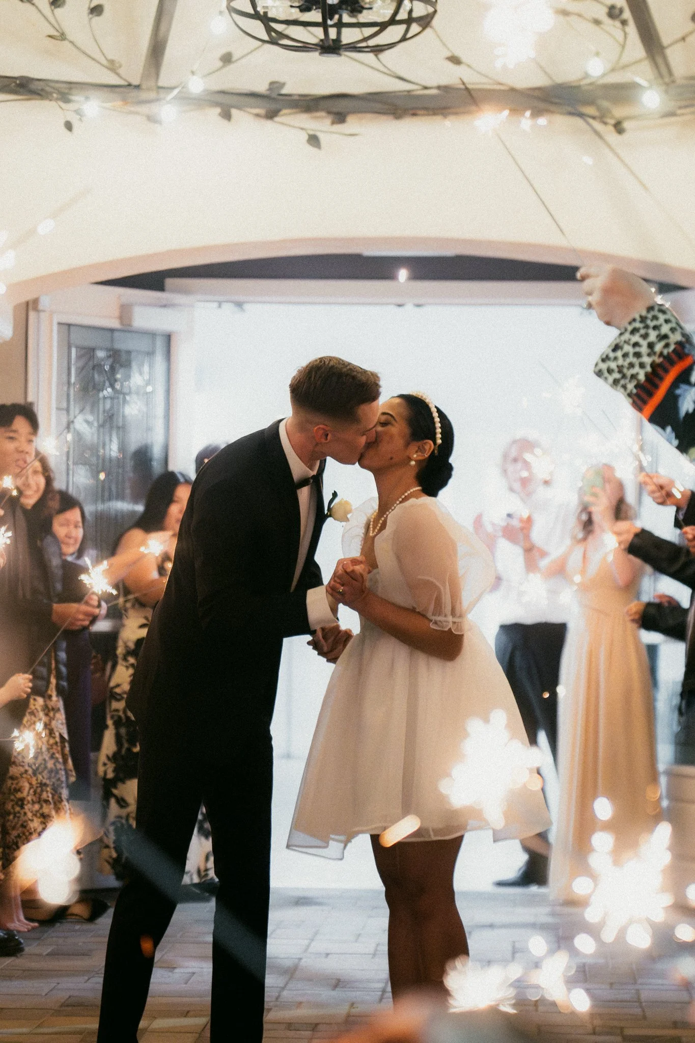A bride and groom kissing during their wedding reception surrounded by guests holding sparklers.