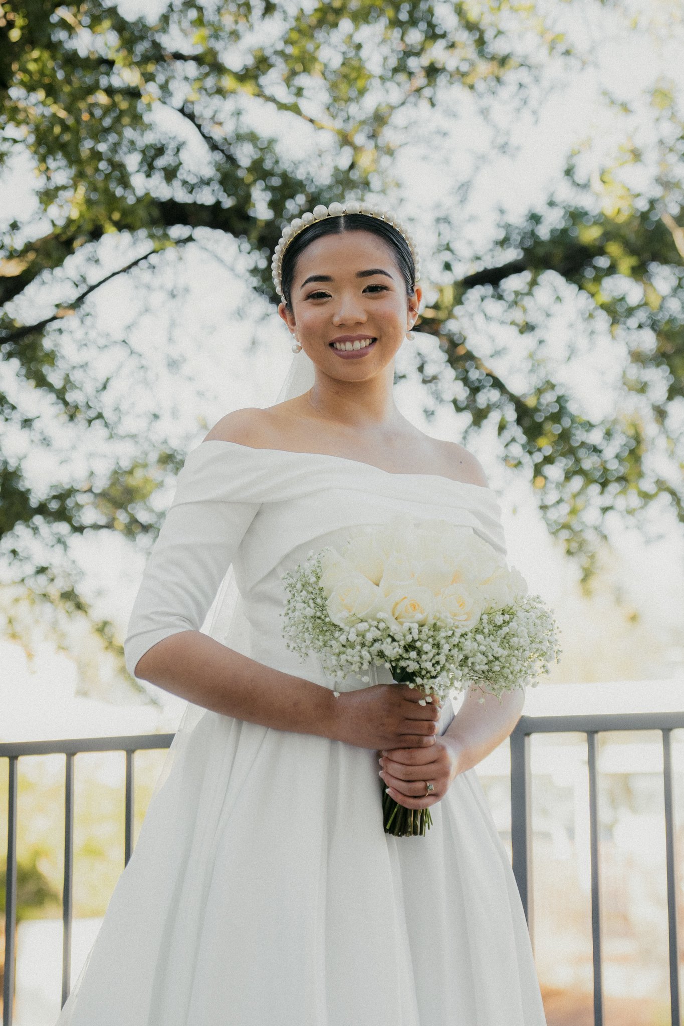 A woman in a white wedding dress holding a bouquet of white roses and baby's breath, standing outdoors with trees and a railing in the background.