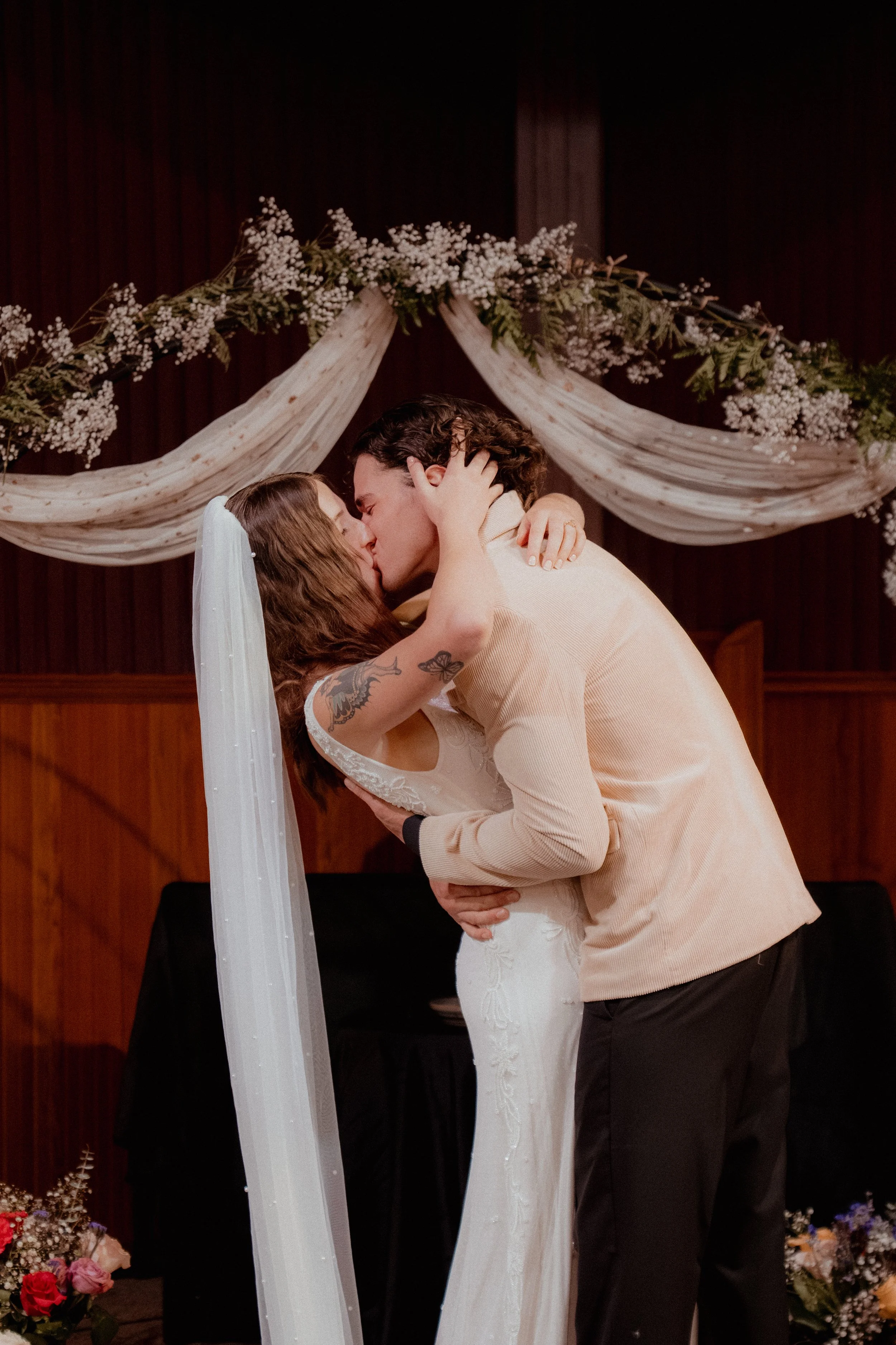 A newlywed couple kissing during their wedding ceremony, with floral decorations and draped fabric in the background.