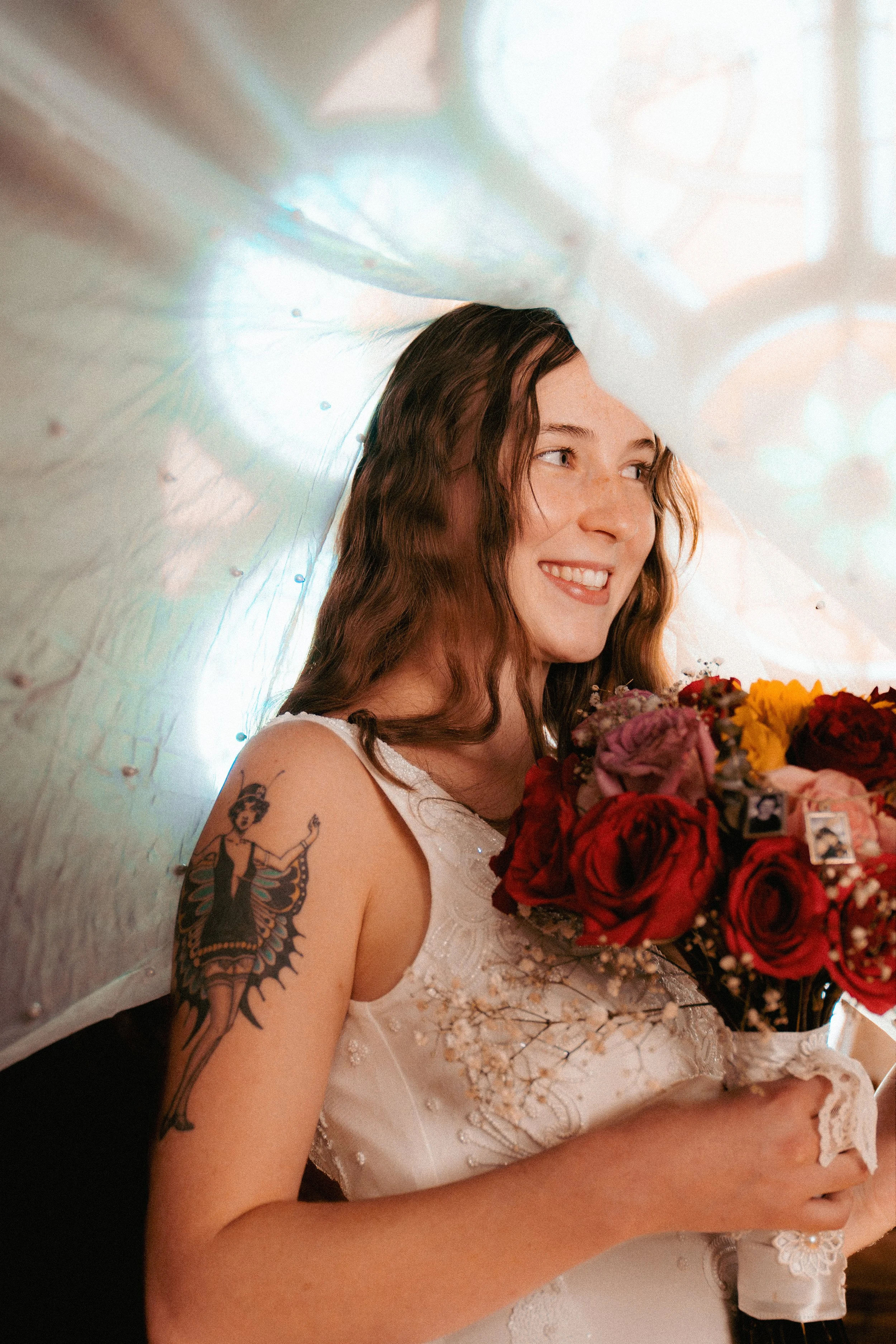 A woman with wavy brown hair holding a bouquet of colorful flowers, smiling. She has a tattoo on her upper arm and is wearing a white dress with lace and embroidery. The background includes a large, iridescent, transparent umbrella.