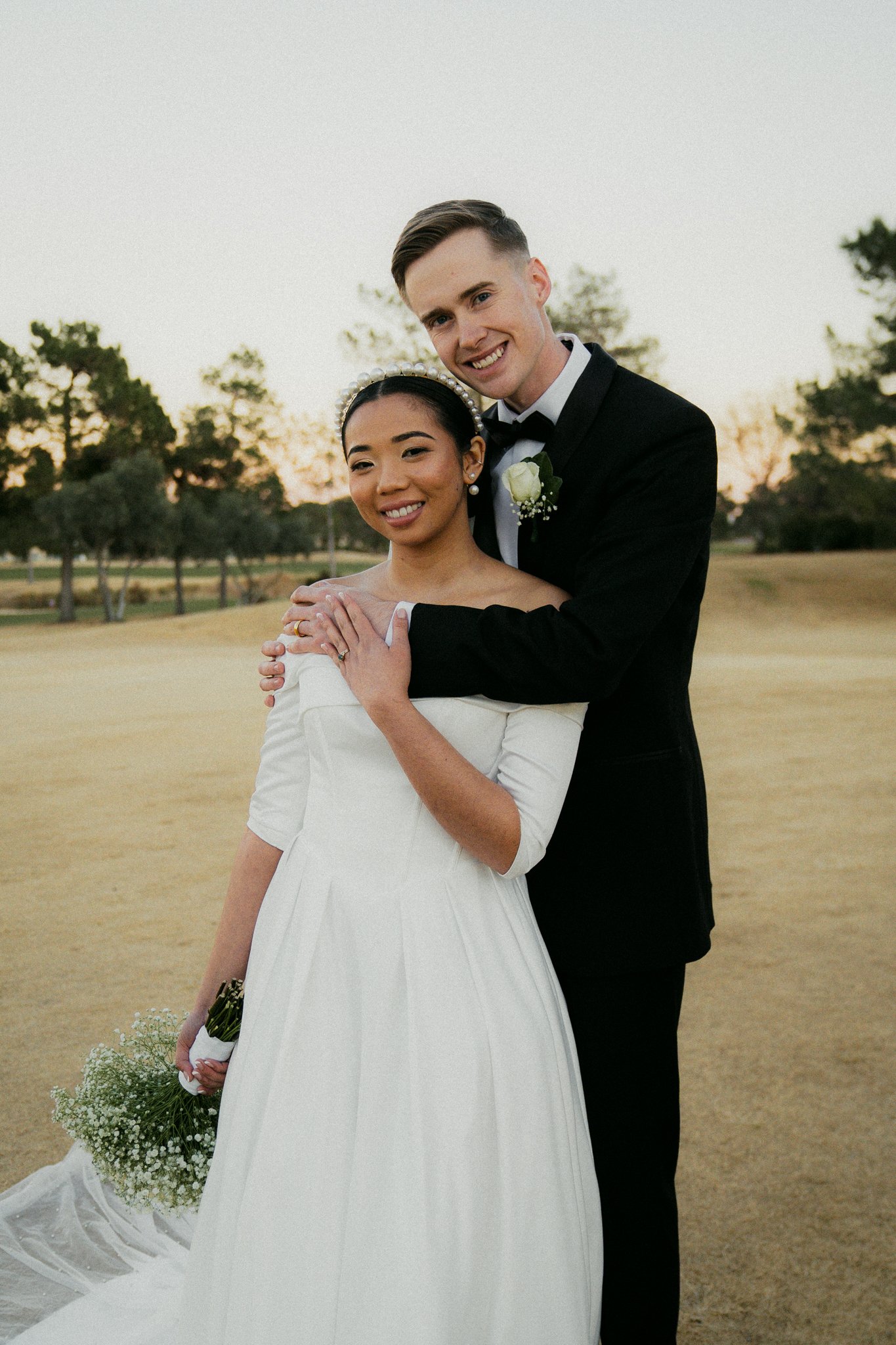 A newlywed couple standing outdoors on a dry, grassy field at sunset, with trees in the background. The bride is wearing a white dress and holding a small bouquet, and the groom is dressed in a black tuxedo with a boutonniere. They are smiling and po