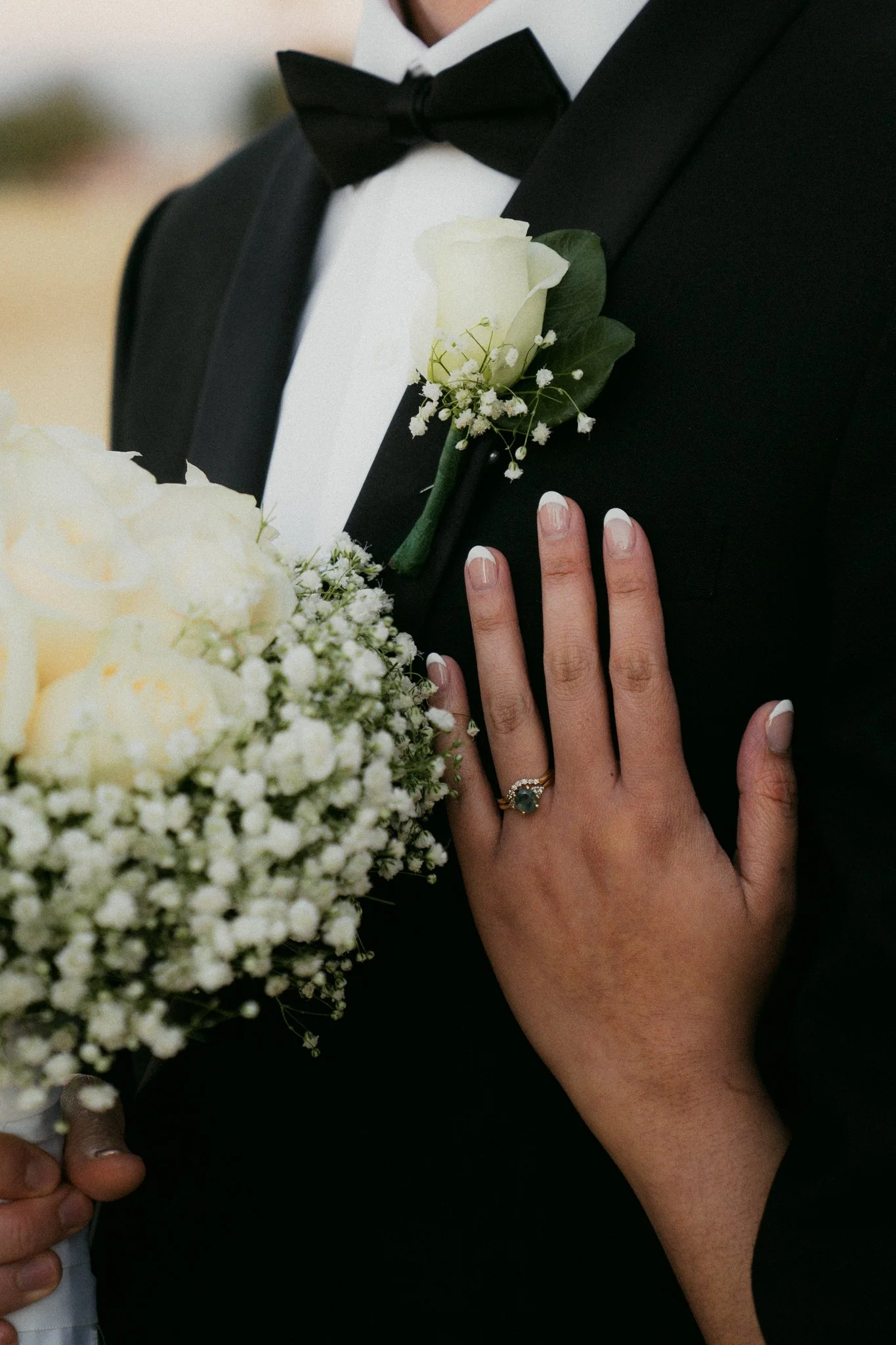 Close-up of a groom in a black tuxedo with a white shirt and black bow tie, holding a bouquet of white roses and baby's breath, with a woman's hand showing a wedding ring resting on his chest.