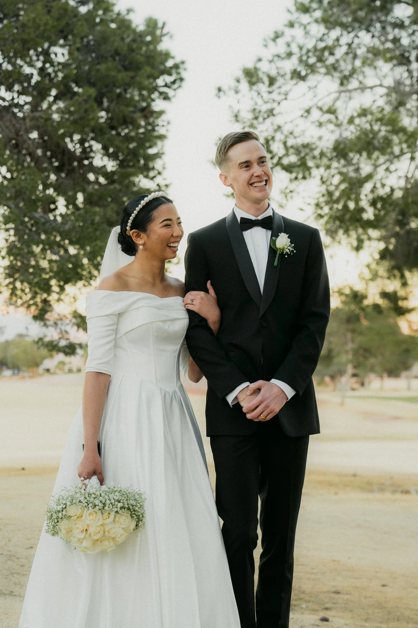 A bride and groom standing outdoors, smiling. The bride is wearing a white wedding dress with puffed sleeves, a pearl headband, and is holding a bouquet of white roses. The groom is dressed in a black tuxedo with a bowtie and white shirt, with a whit