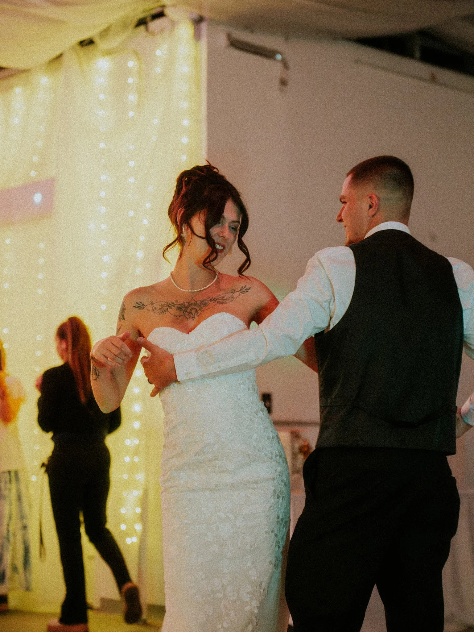 A bride and groom dancing at their wedding reception. The bride is wearing a strapless, lace wedding dress and has dark, curly hair. The groom is dressed in a white shirt with a black vest and pants. They are smiling and looking at each other, with a
