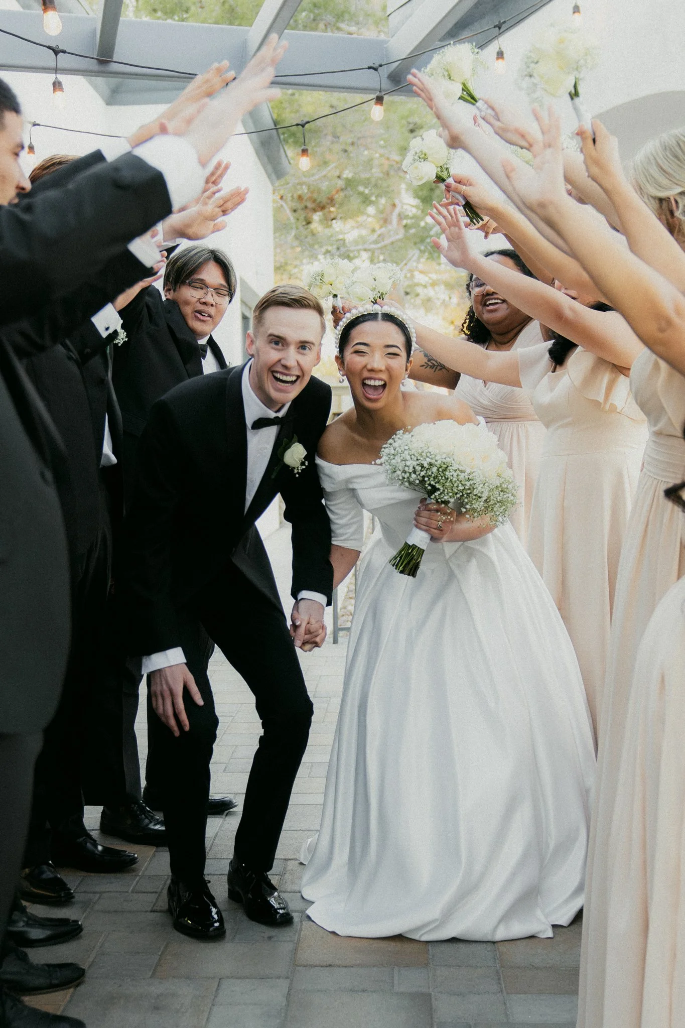 Happy bride and groom holding hands, surrounded by smiling bridesmaids and groomsmen, celebrating during wedding exit with raised flower bouquets and joyful expressions.