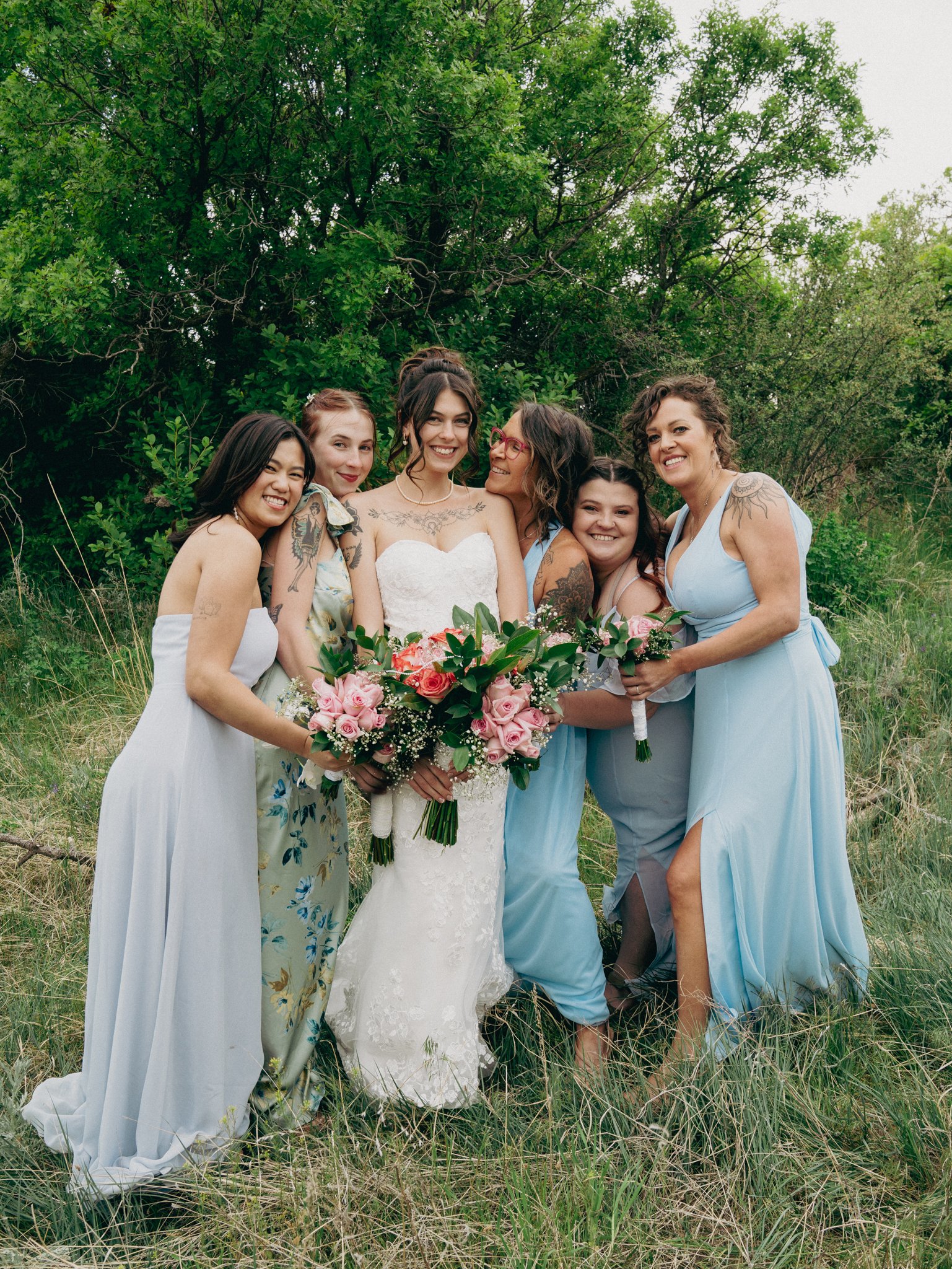 A group of six women, including a bride in a white wedding dress, standing outdoors on grass with green trees in the background, smiling and holding bouquets of pink roses and greenery.