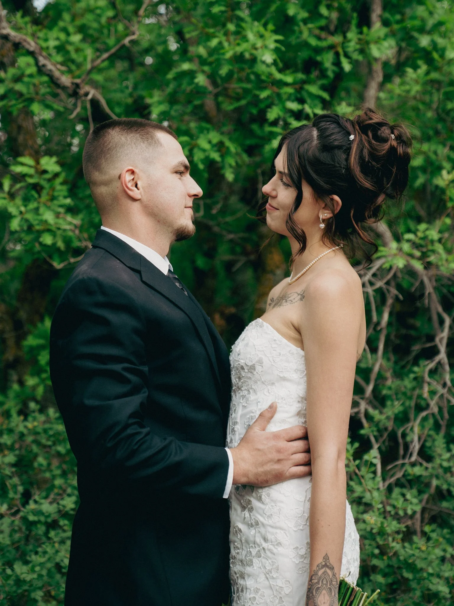 A newlywed couple standing close together outdoors, facing each other with their eyes closed, surrounded by lush green foliage.