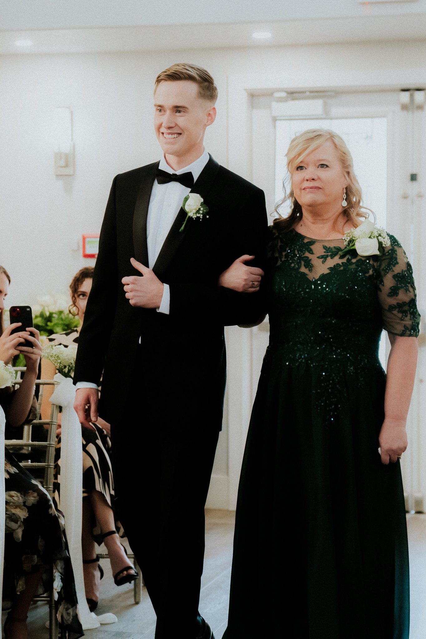 A young man in a tuxedo and a woman in a black dress walking down an aisle during a wedding ceremony, with guests watching and taking photos.