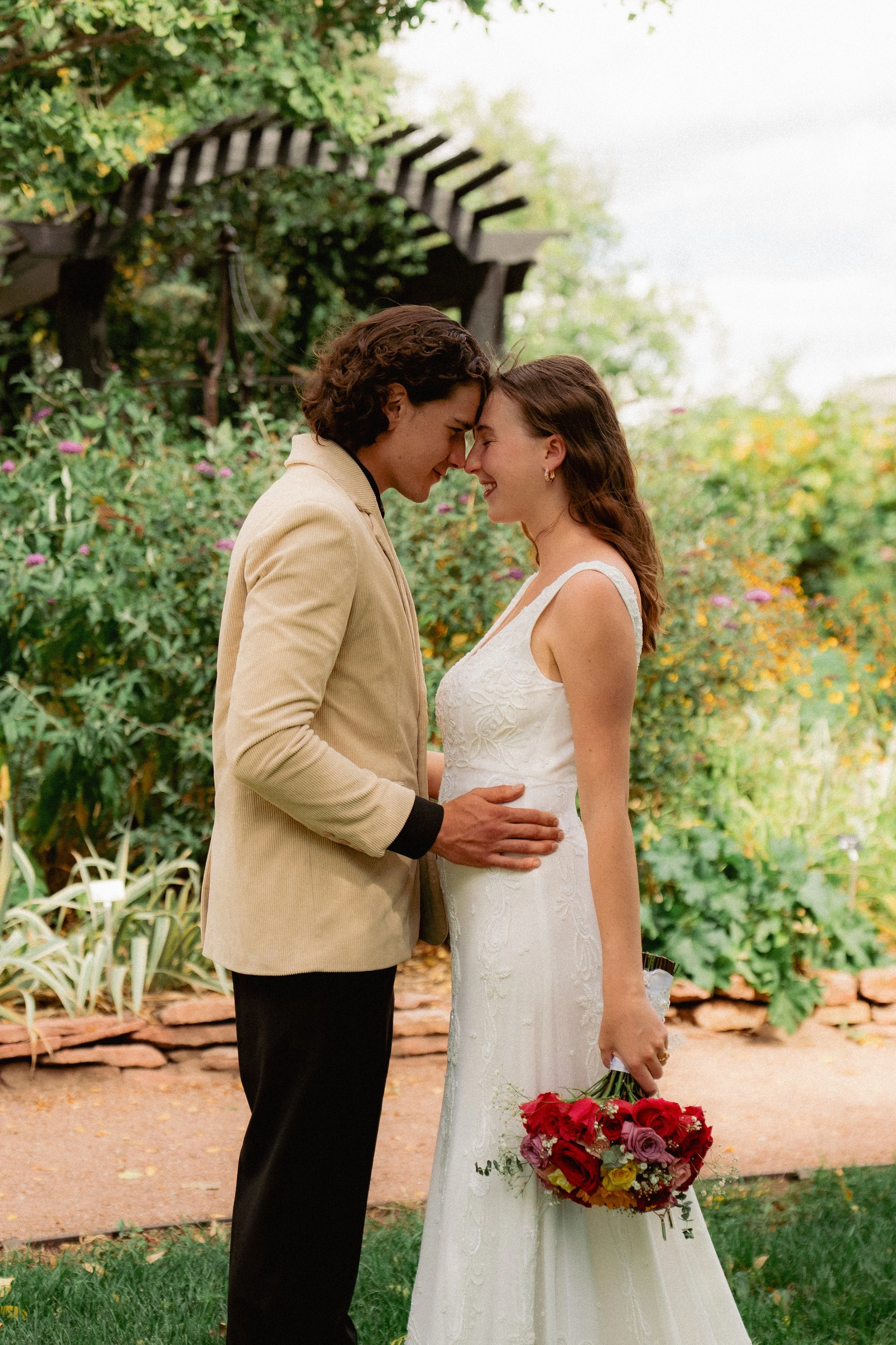 A couple touching foreheads and smiling during a wedding or romantic photoshoot outdoors, with a woman holding a bouquet of red and yellow flowers.