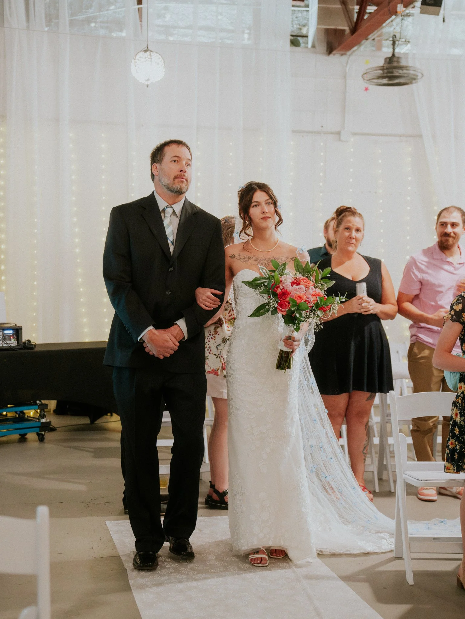 A bride and groom walking down the aisle during their wedding ceremony, with friends and family watching in a decorated venue with white drapes and string lights.
