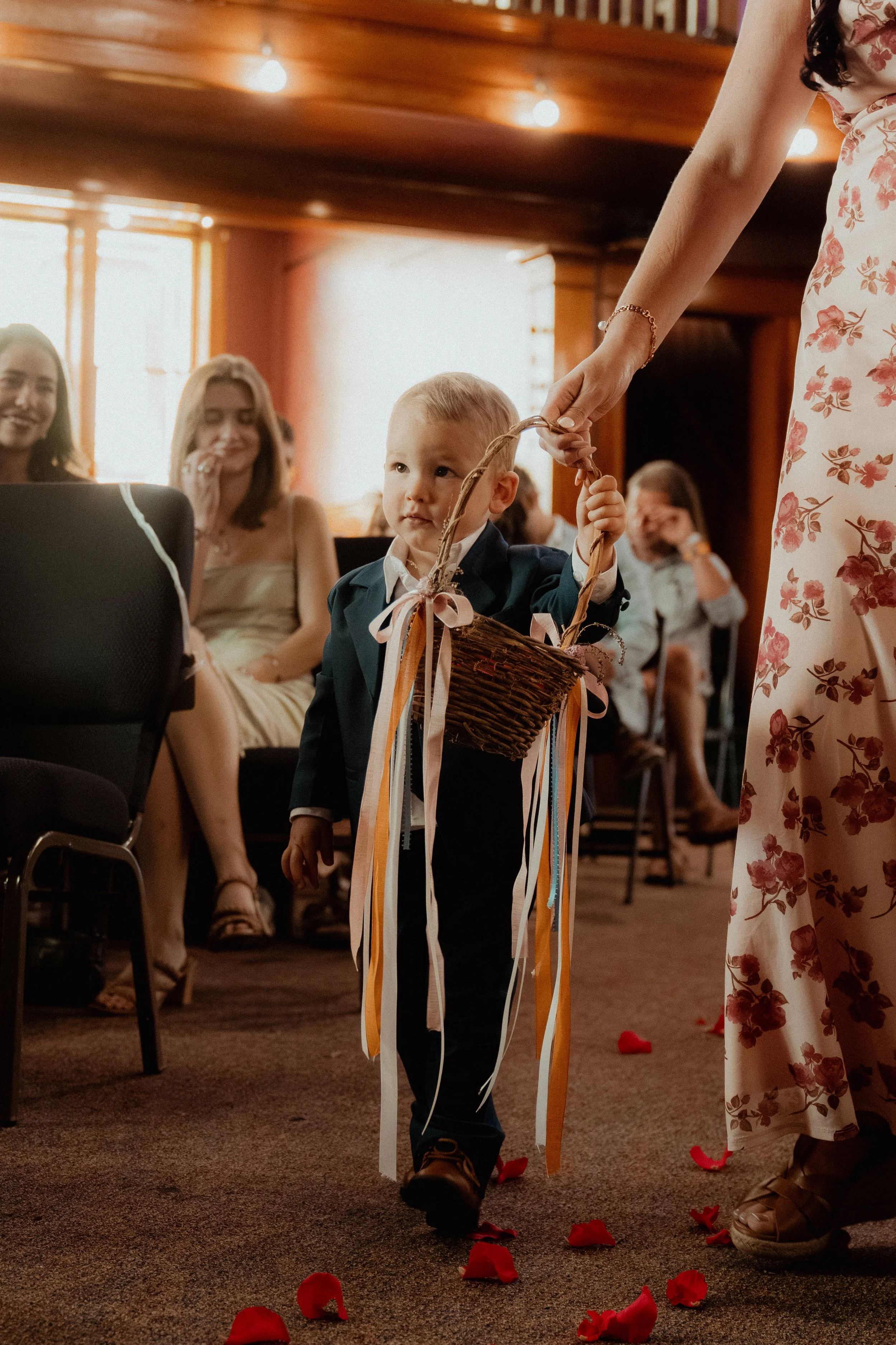 A young boy dressed in a suit is carrying a basket decorated with ribbons at a wedding or celebration, walking barefoot on a carpeted floor strewn with rose petals, accompanied by an adult woman in a floral dress, with guests seated and smiling in th
