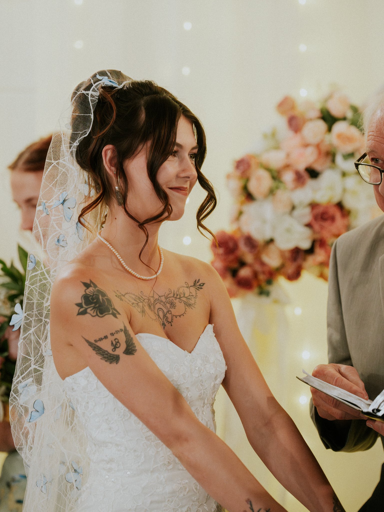 A bride in a white wedding dress with tattoos on her arms and chest, wearing a pearl necklace and veil, smiling during her wedding ceremony.
