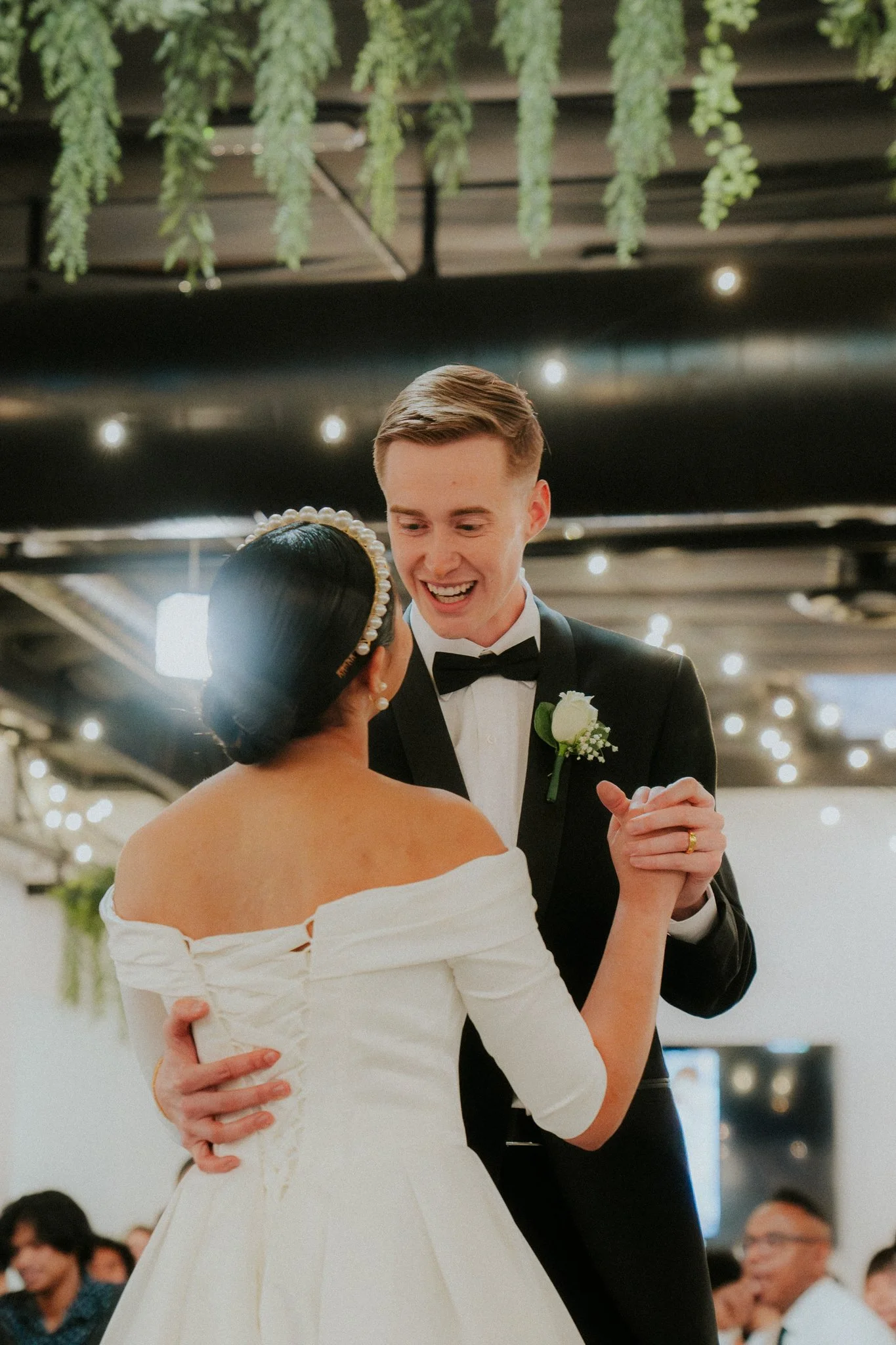 A bride and groom dancing at their wedding reception. The bride is wearing an off-shoulder white gown with a pearl headband, and the groom is dressed in a black tuxedo with a bow tie and a white boutonniere. They are smiling and holding hands as they