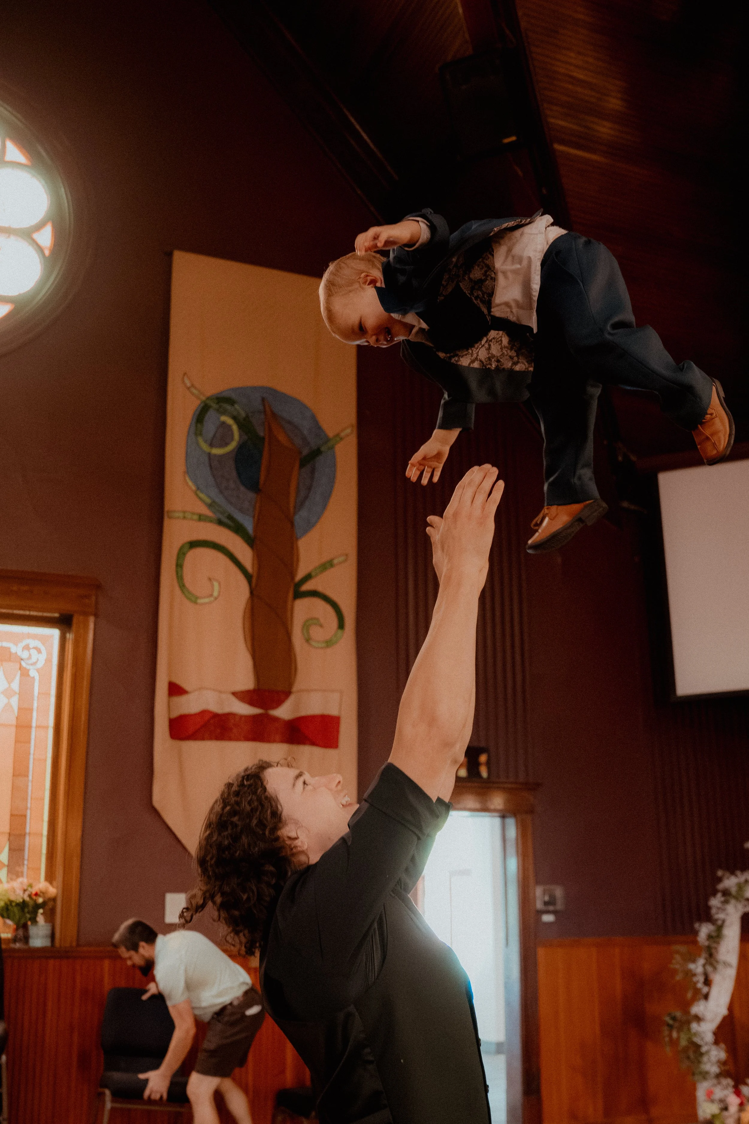 A woman with curly hair smiling and reaching up as she lifts a young girl into the air inside a room with wooden walls and colorful artwork hanging on the wall.