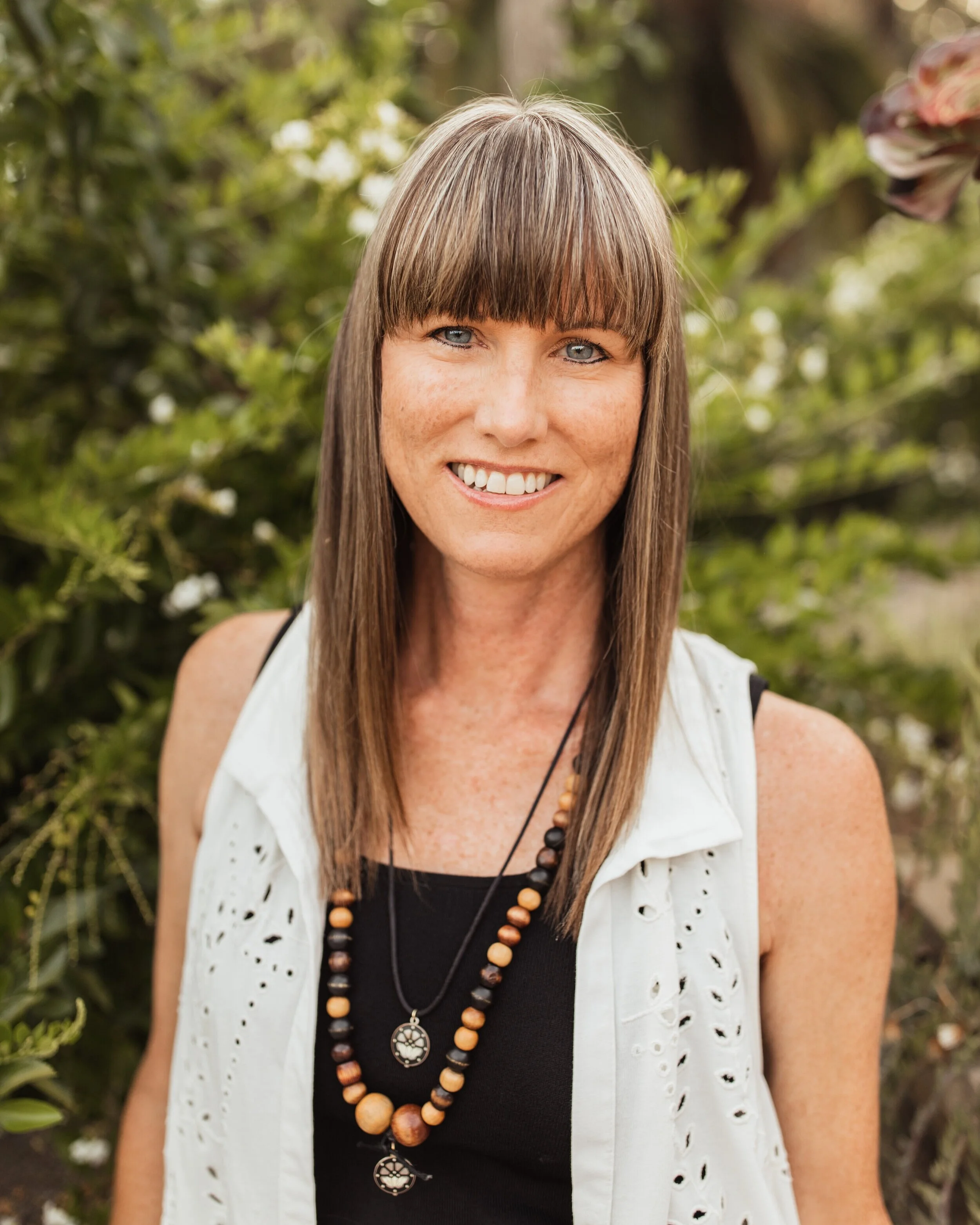 A woman with brown hair wearing a sleeveless white vest, a black top, and layered necklaces.