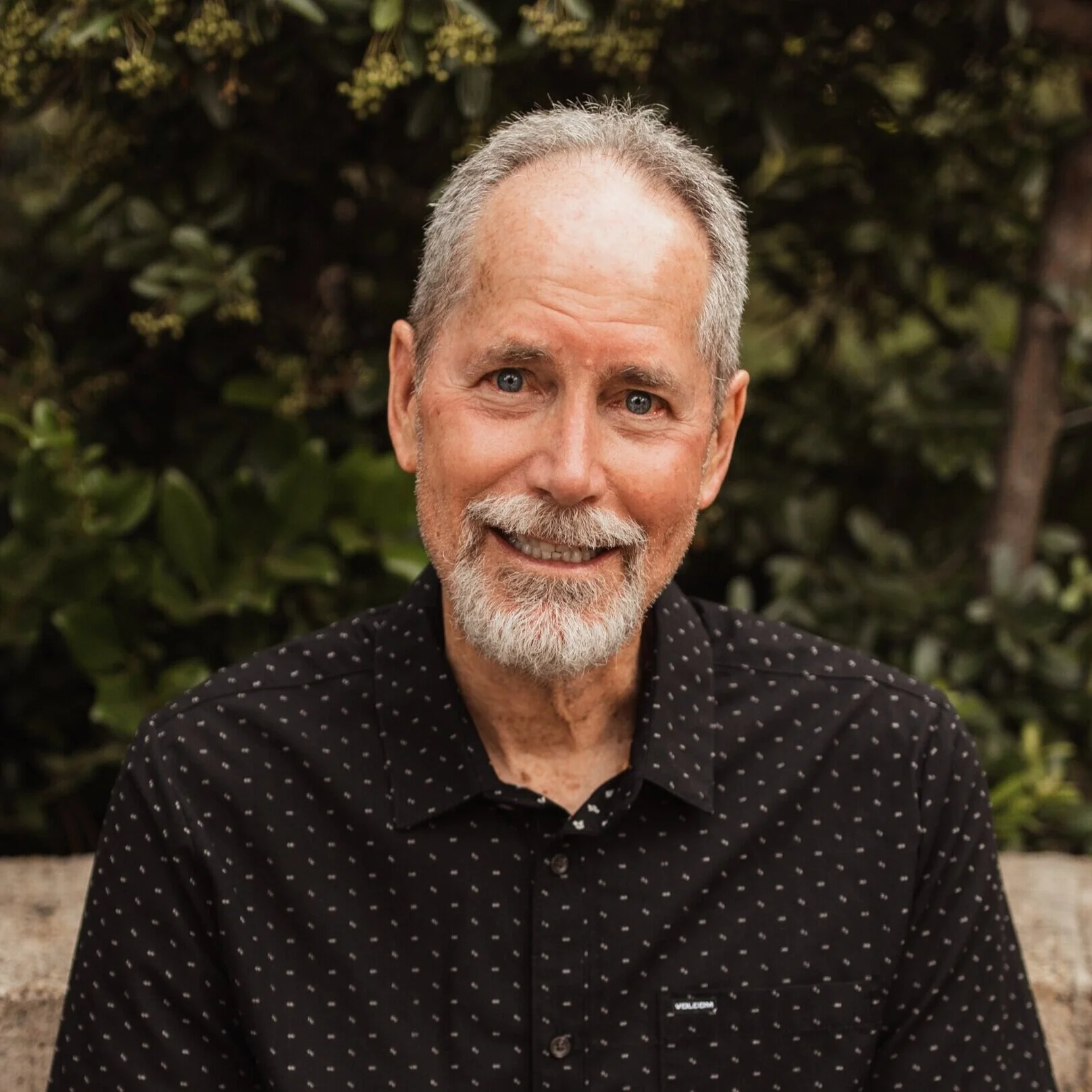 A smiling man with a beard and mustache, wearing a black short-sleeved button-up shirt with white dots, sitting outdoors with green foliage in the background.