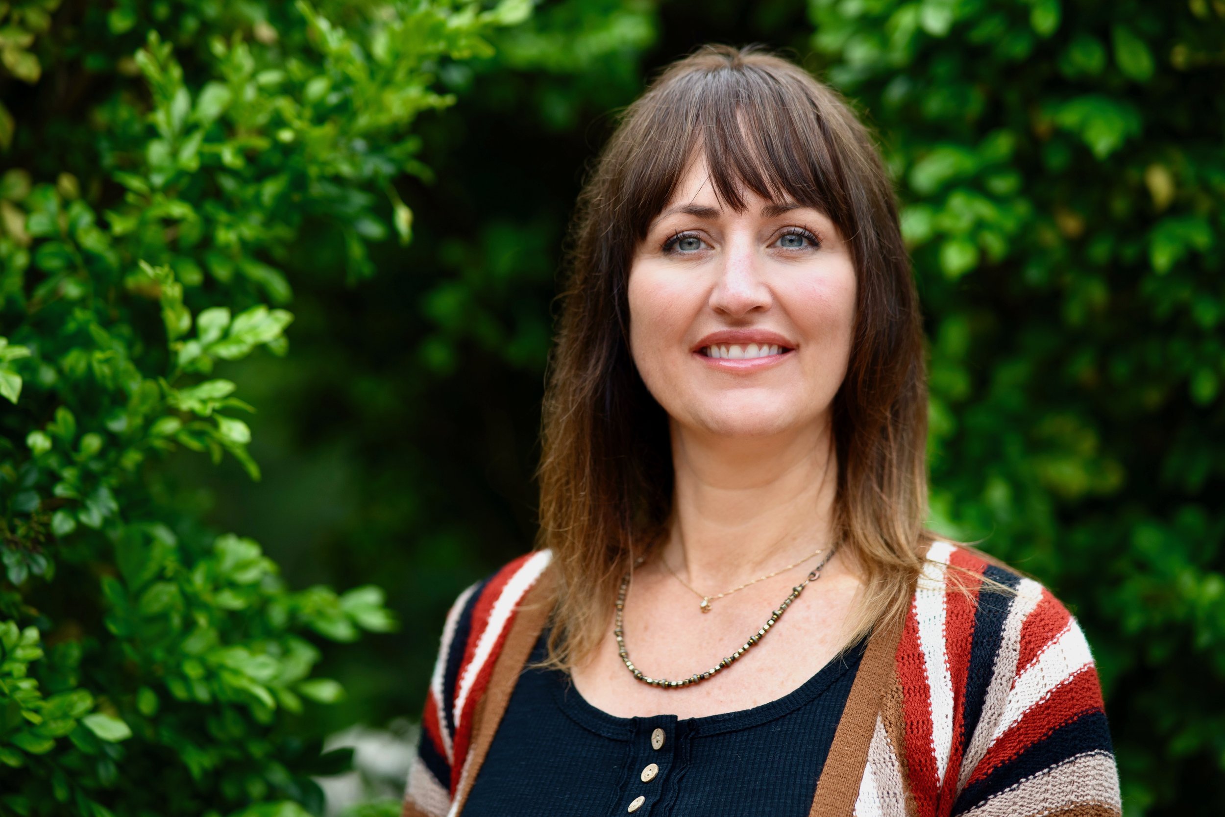 A smiling middle aged woman with long brown hair standing outdoors among green foliage, wearing a multicolored sweater (white, black, red, brown), black top, and a beaded necklace. 