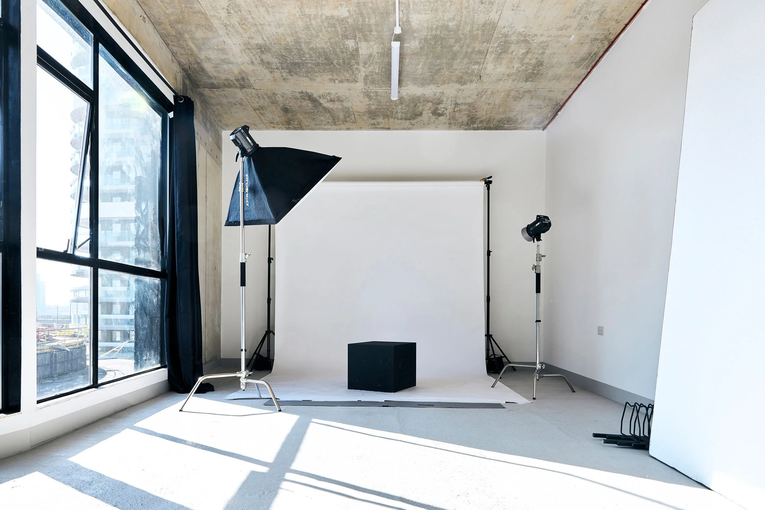 Nocturna Photography (Studio Monox) based in Royal Victoria DOcks (London). The picture of the studio shows the studio with a white backdrop, studio lights, and black cube on the floor near large window with city view, sunlight casting shadows.