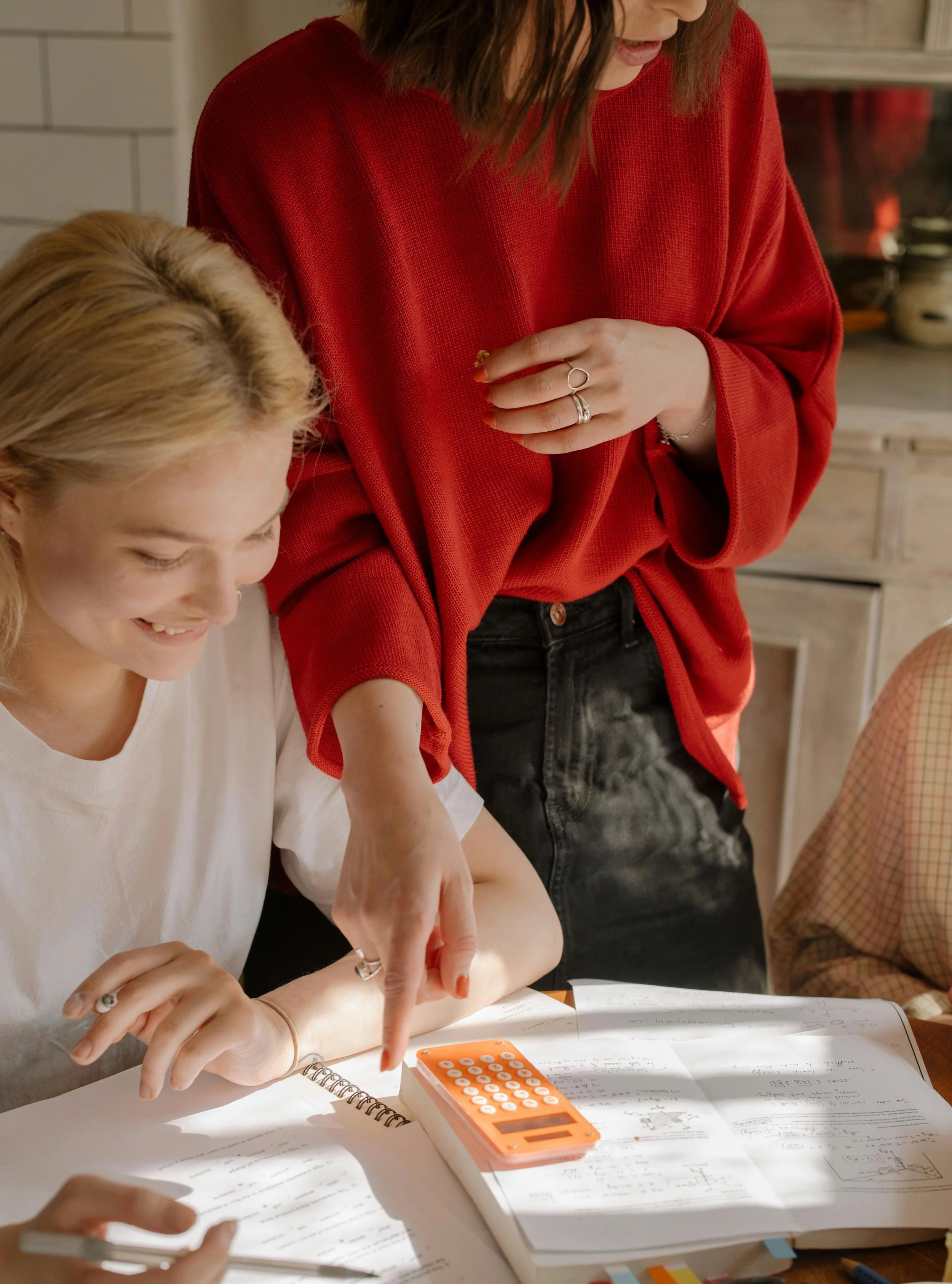 Parent helping a teen with homework and a calculator at the kitchen table, building life skills for young adults like planning and problem-solving.