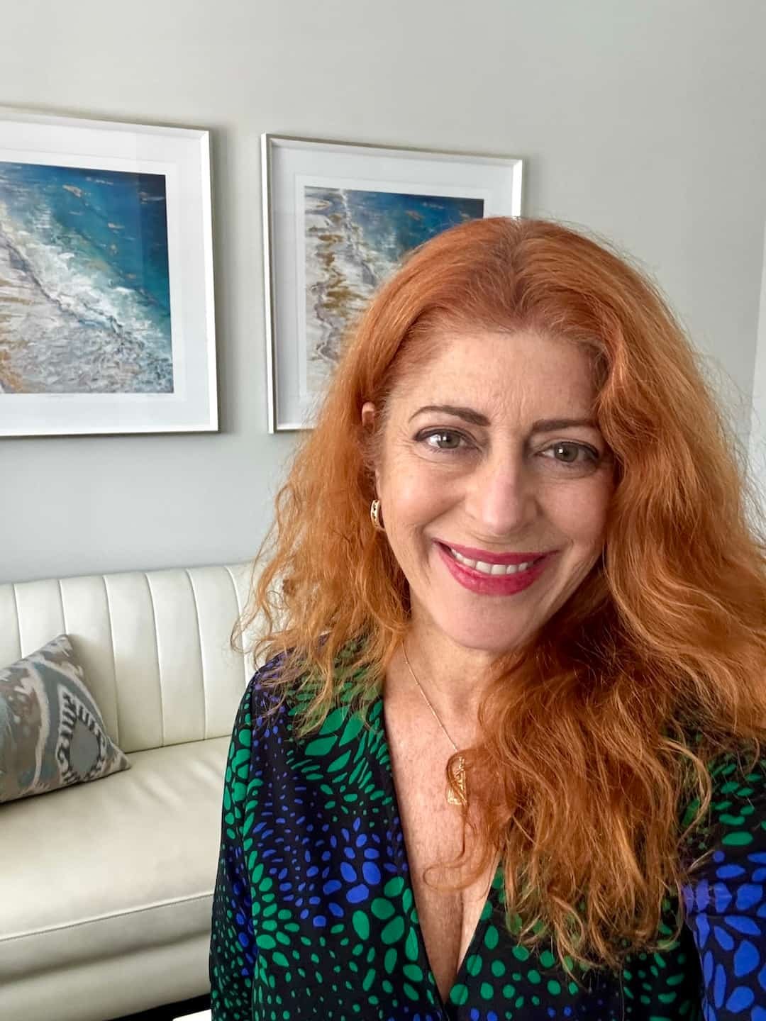 woman with red hair wearing a blue and green printed top in front of a white couch