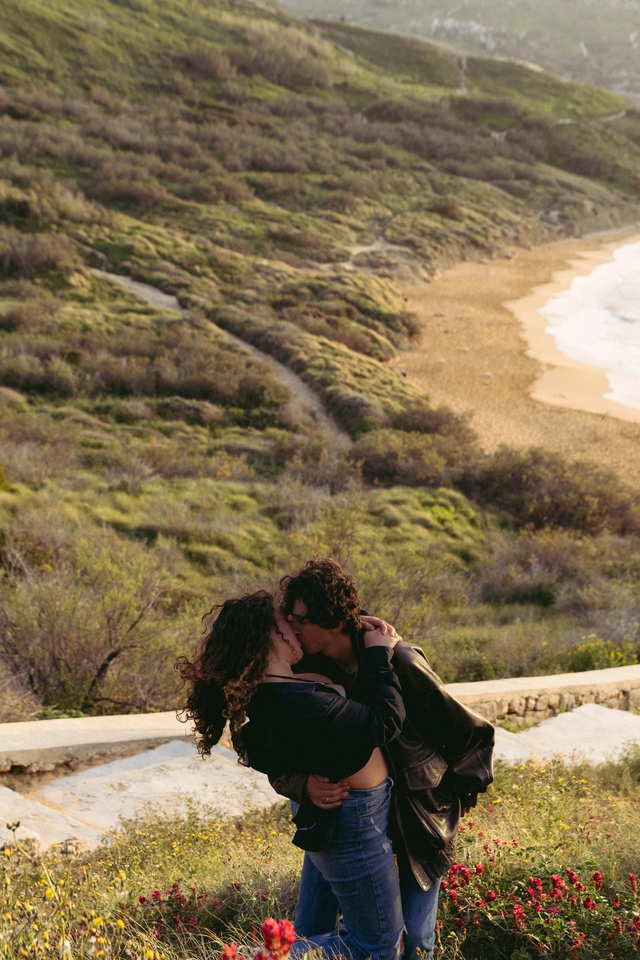 Couple kissing near a scenic coastline with greenery and a sandy beach.
