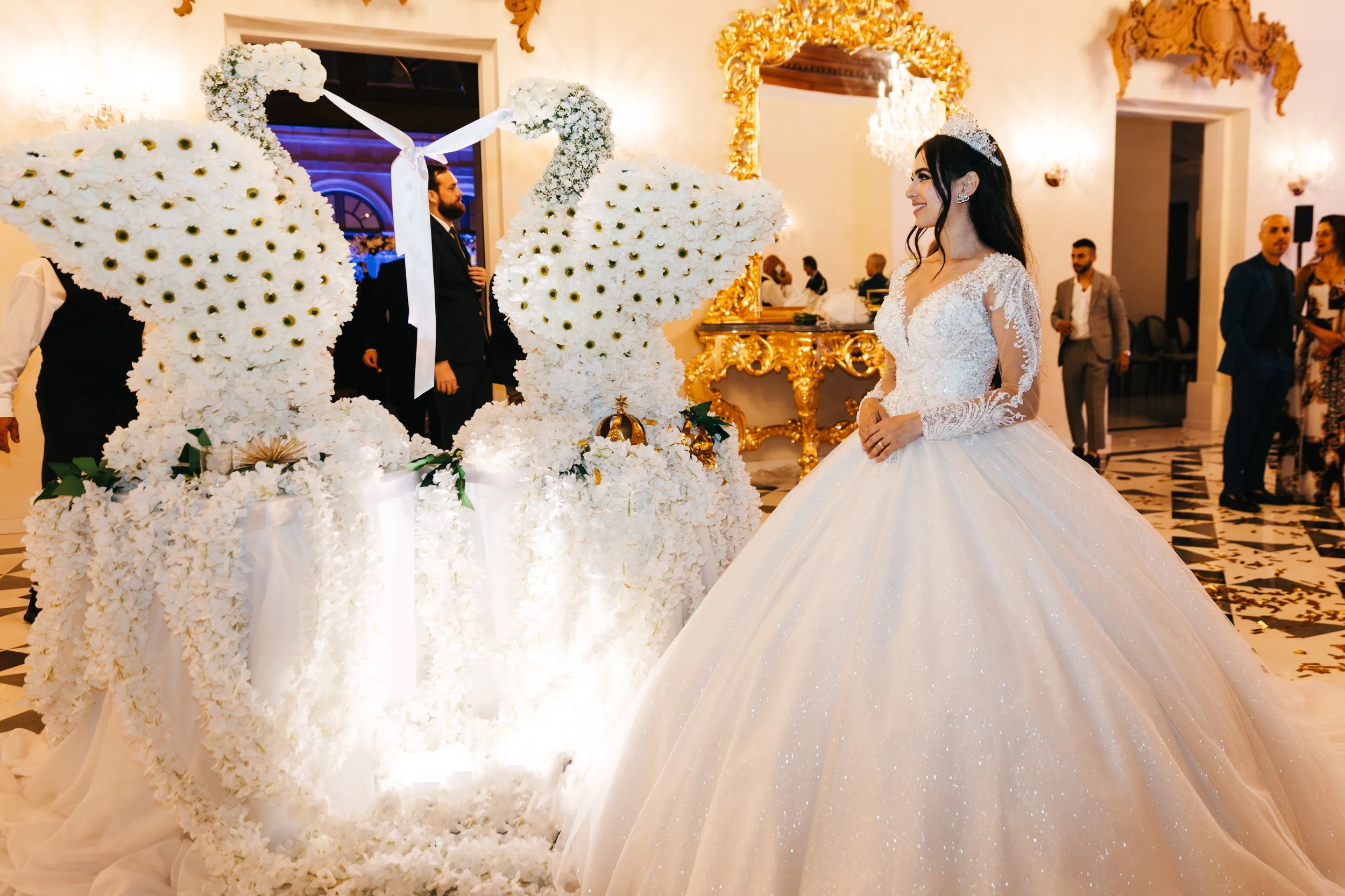 Bride in a wedding dress next to two decorative floral peacocks with guests in the background.
