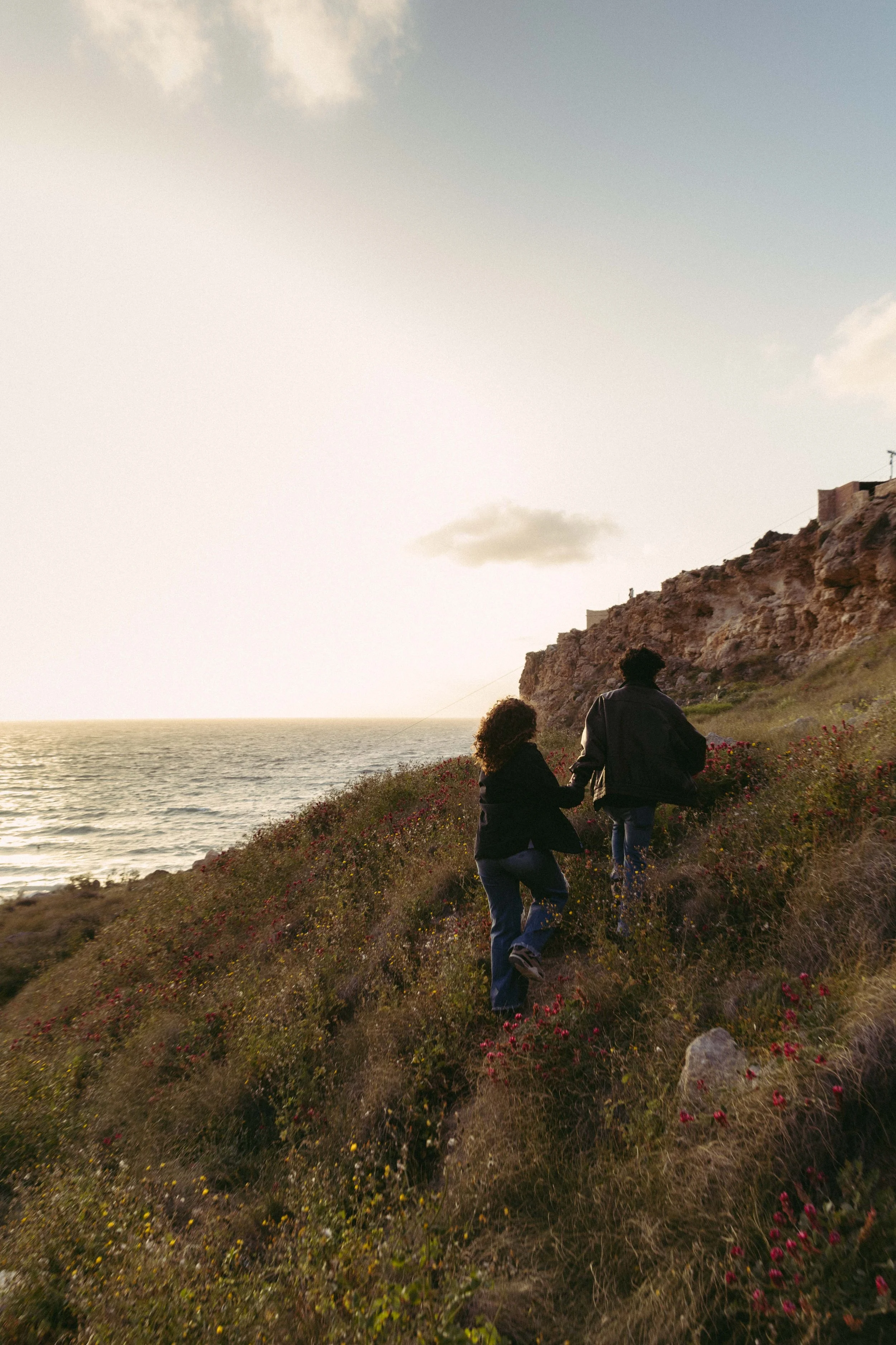 Two people walking uphill near a cliff by the sea during sunset.