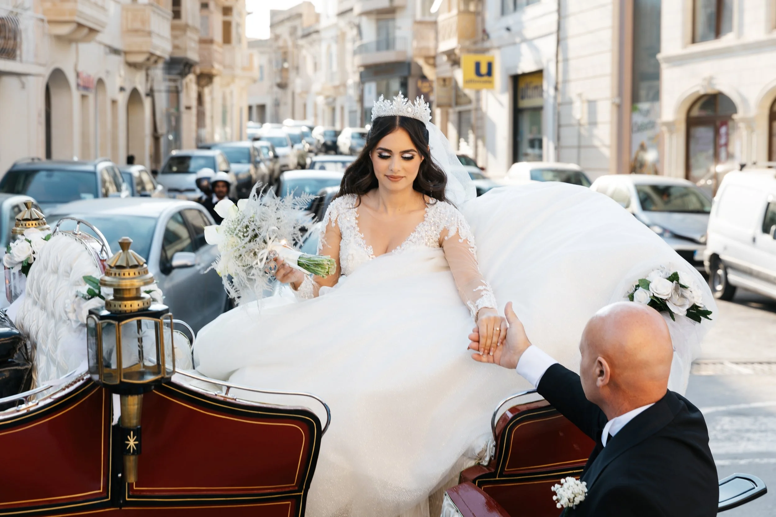 Bride in a white gown holding a bouquet, sitting in a horse-drawn carriage, with a man dressed formally reaching out to her, on a city street.