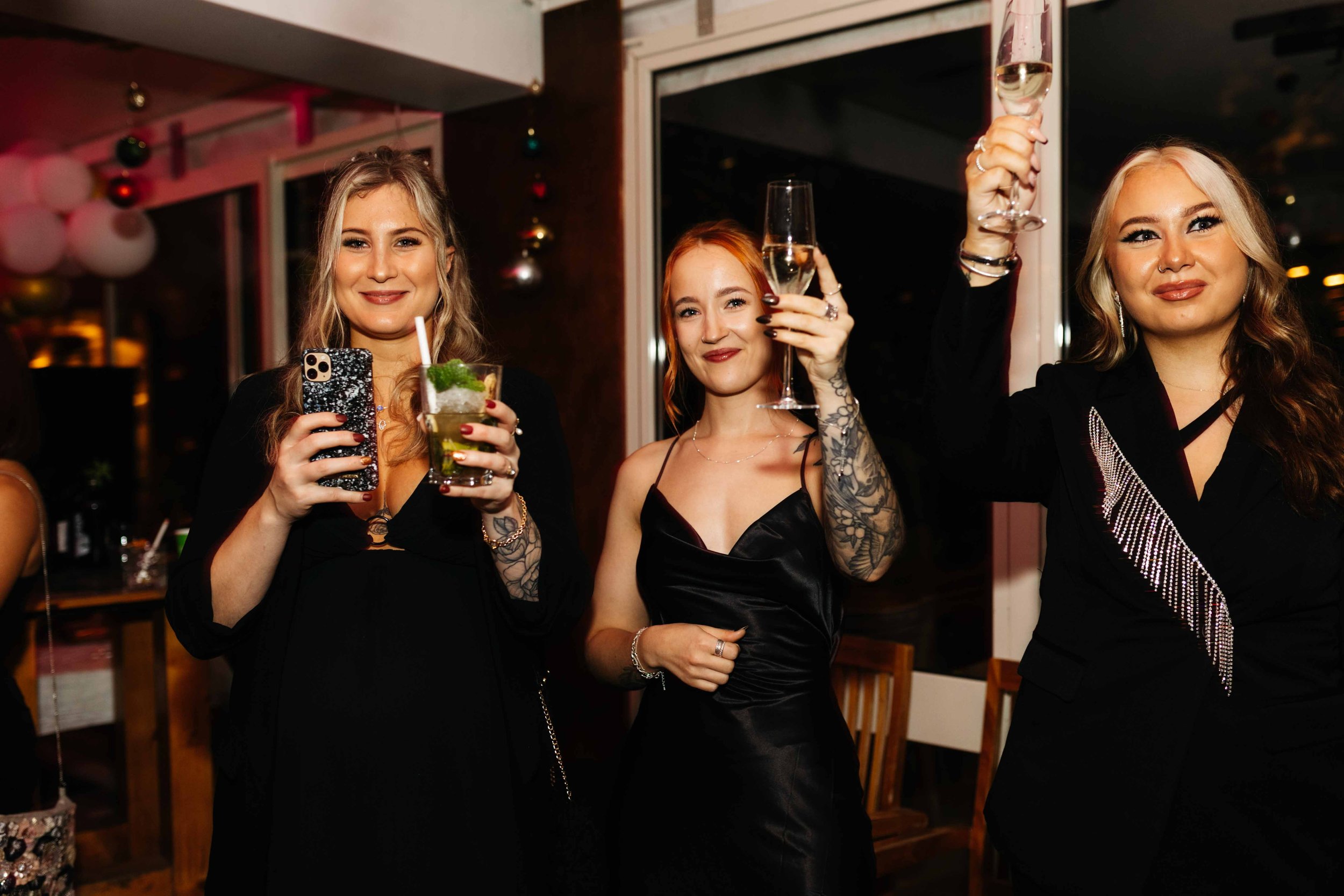 Three women in a celebratory setting, holding drinks, dressed in evening attire.