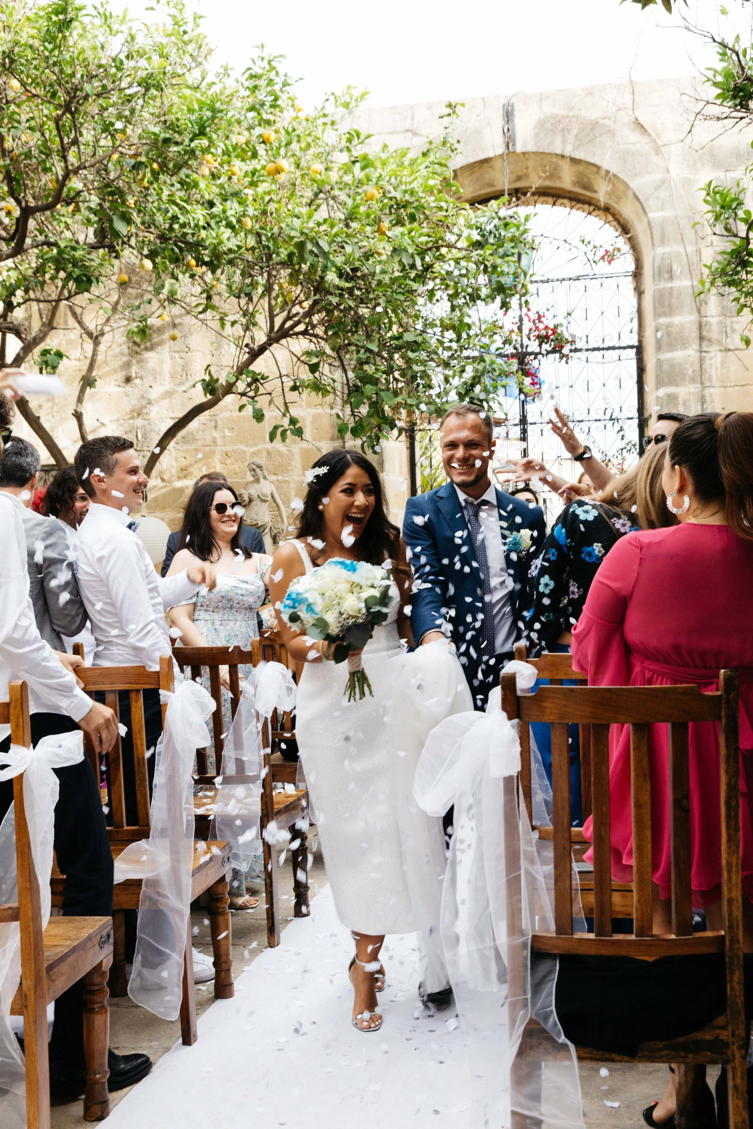 Bride and groom exiting wedding ceremony under flower petals, surrounded by guests, outdoor setting with wooden chairs and greenery.