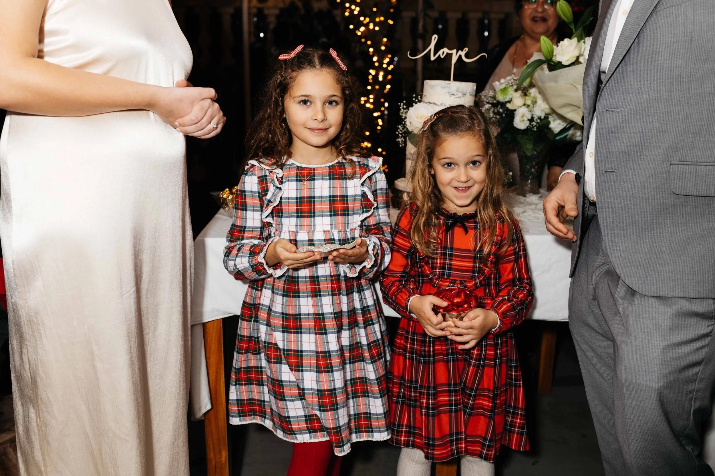 Two young girls in plaid dresses holding small gifts, standing with adults in elegant attire at a festive event with a table and cake in the background.