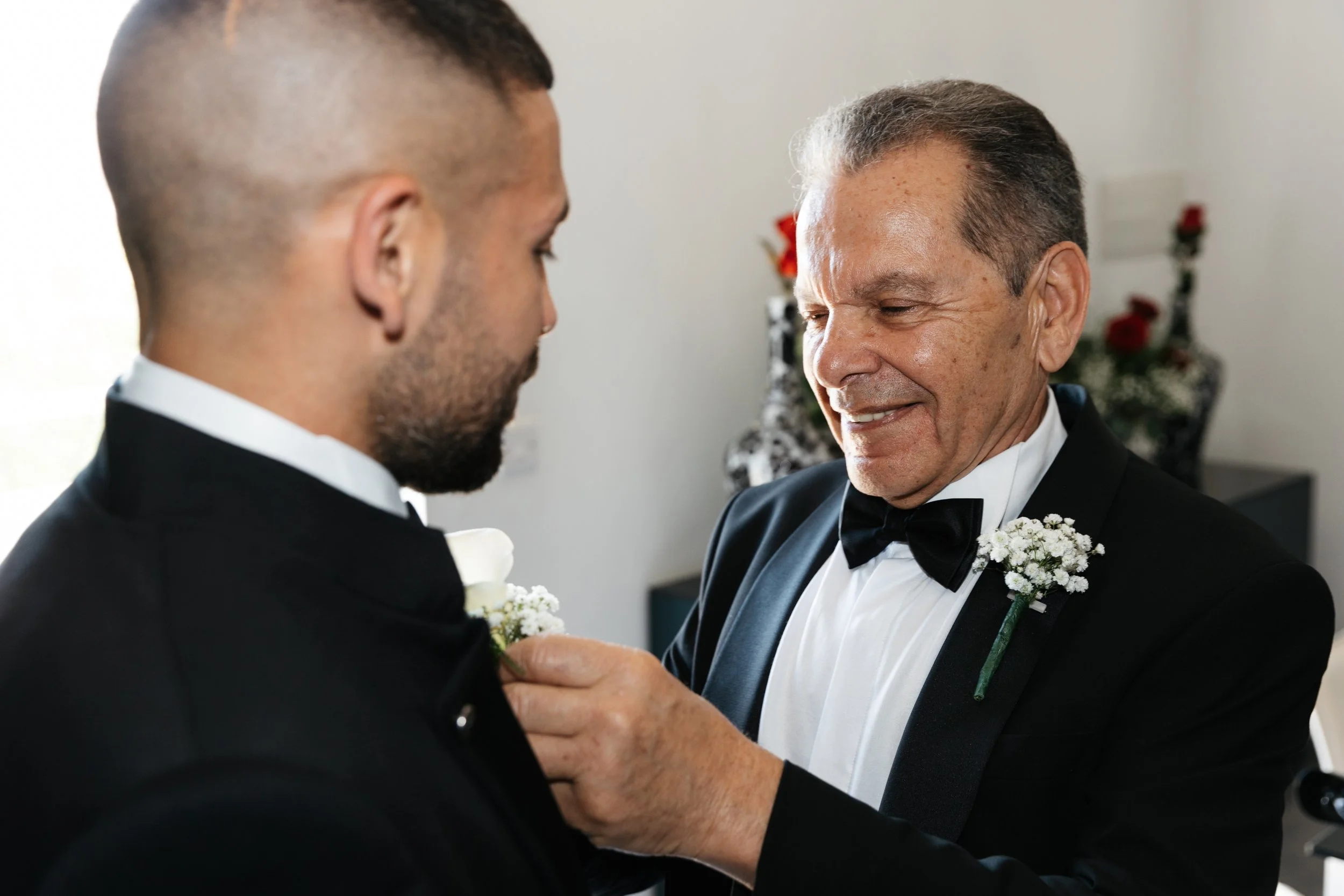 An elderly man in a tuxedo pinning a boutonniere on a young man's lapel, smiling, in a formal setting.